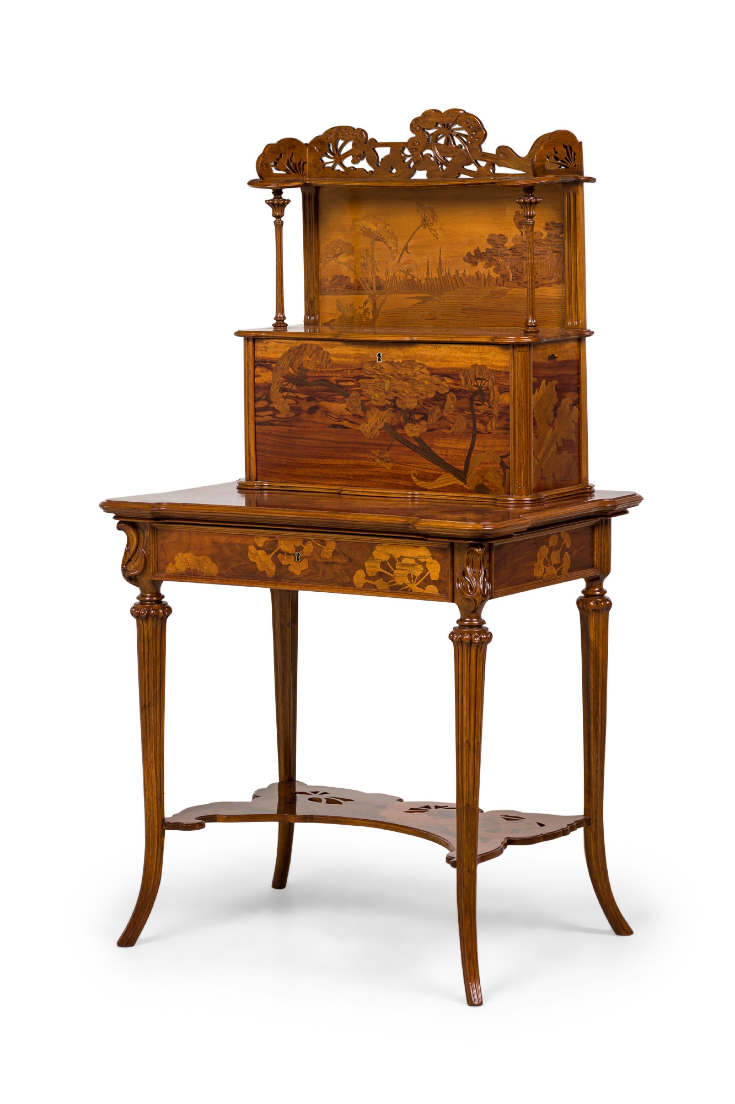 French Art Nouveau writing desk featuring an open upper section with a marquetry inlaid pasteral scene back panel under a carved floral crest supported by two fluted & carved columns over a drop front cabinet having a floral marquetry cabinet