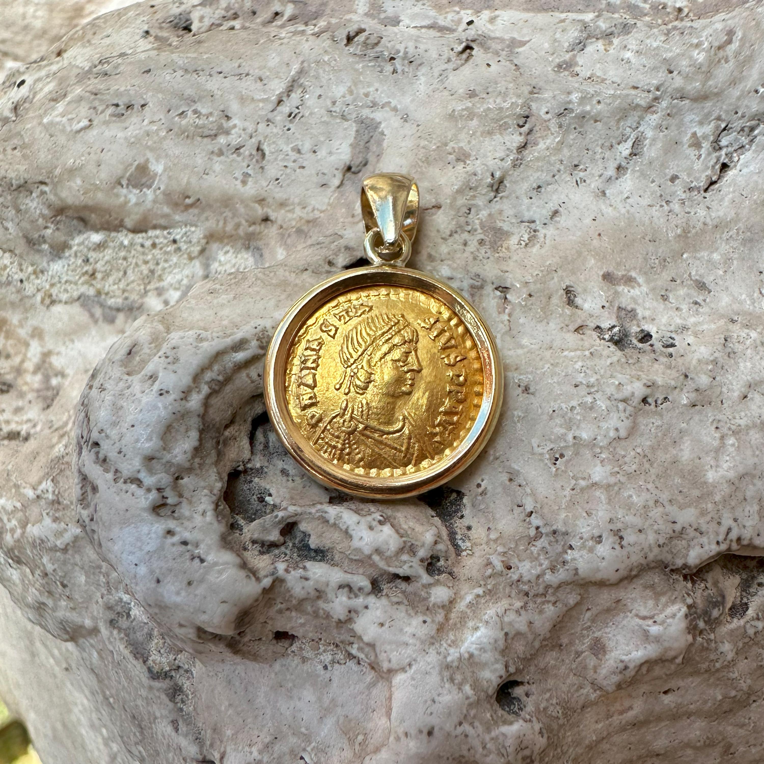 This exquisite 18Kt Gold pendant houses a Byzantine Tremissis from the 5th century AD, featuring the Emperor Anastasius I. On the front, we see his helmeted and cuirassed bust facing forward, holding a spear and shield, symbolizing his military