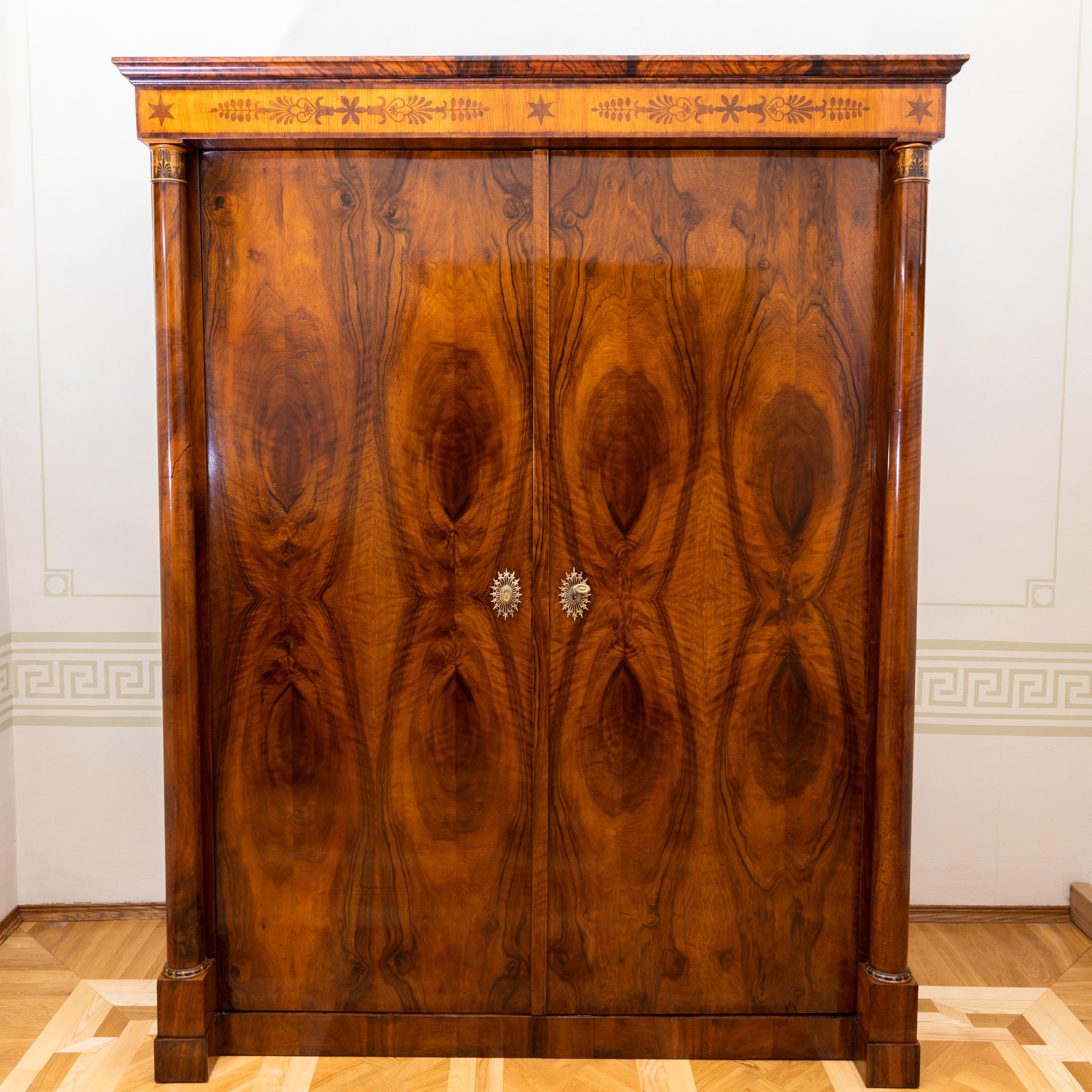Empire cabinet with two doors, in solid and veneered walnut. The strict corpus is flanked by columns with floral ink painting on the capitals and bases. The straight profiled cornice with inlayed ornamental band with palmettes and stars completes