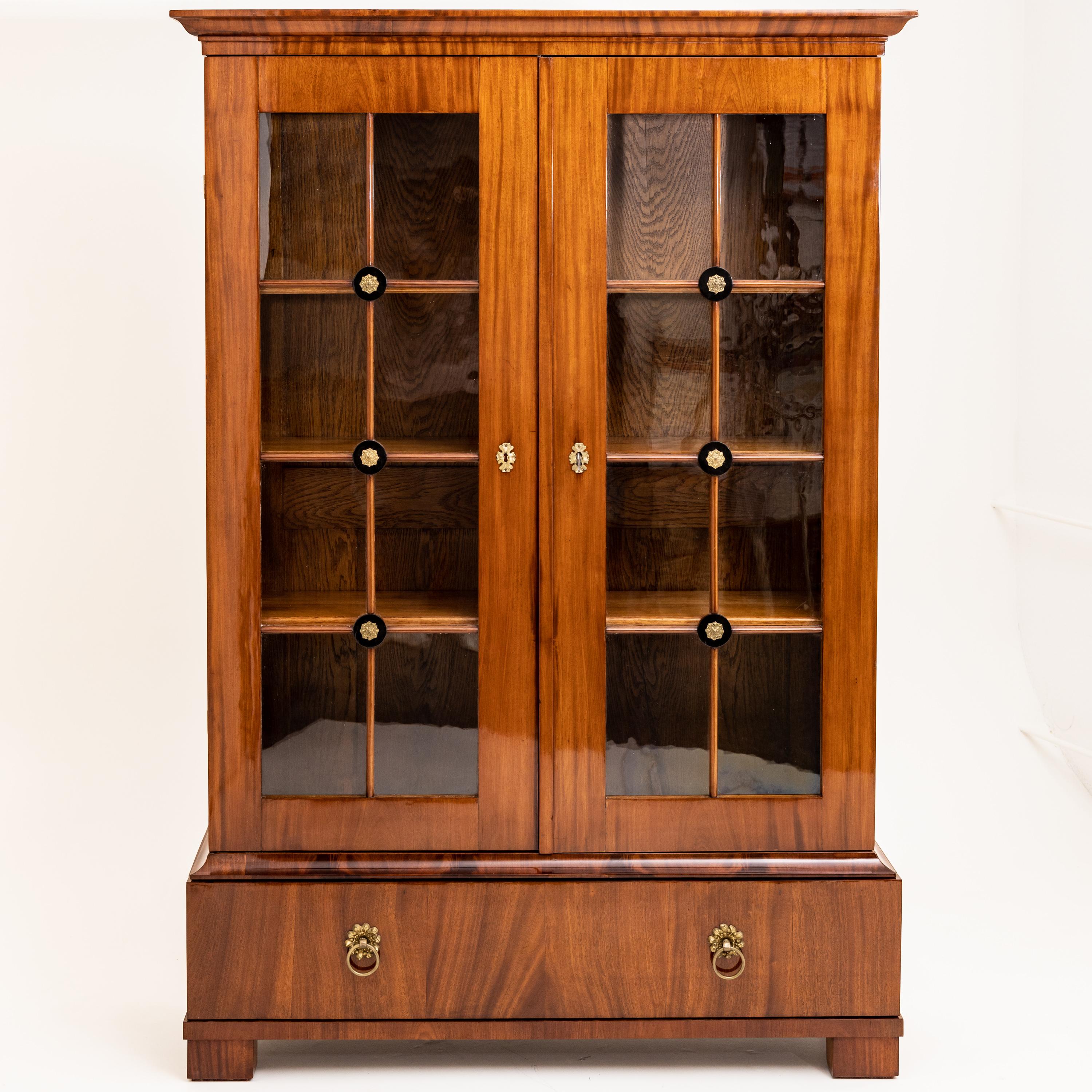 Two-door bookcase on a high base with one drawer and ring-shaped handles in bronze. The glazing of the doors is grated and decorated with fire-gilded bronze rosettes at the crossing points. The interior consists of three shelves adjustable in