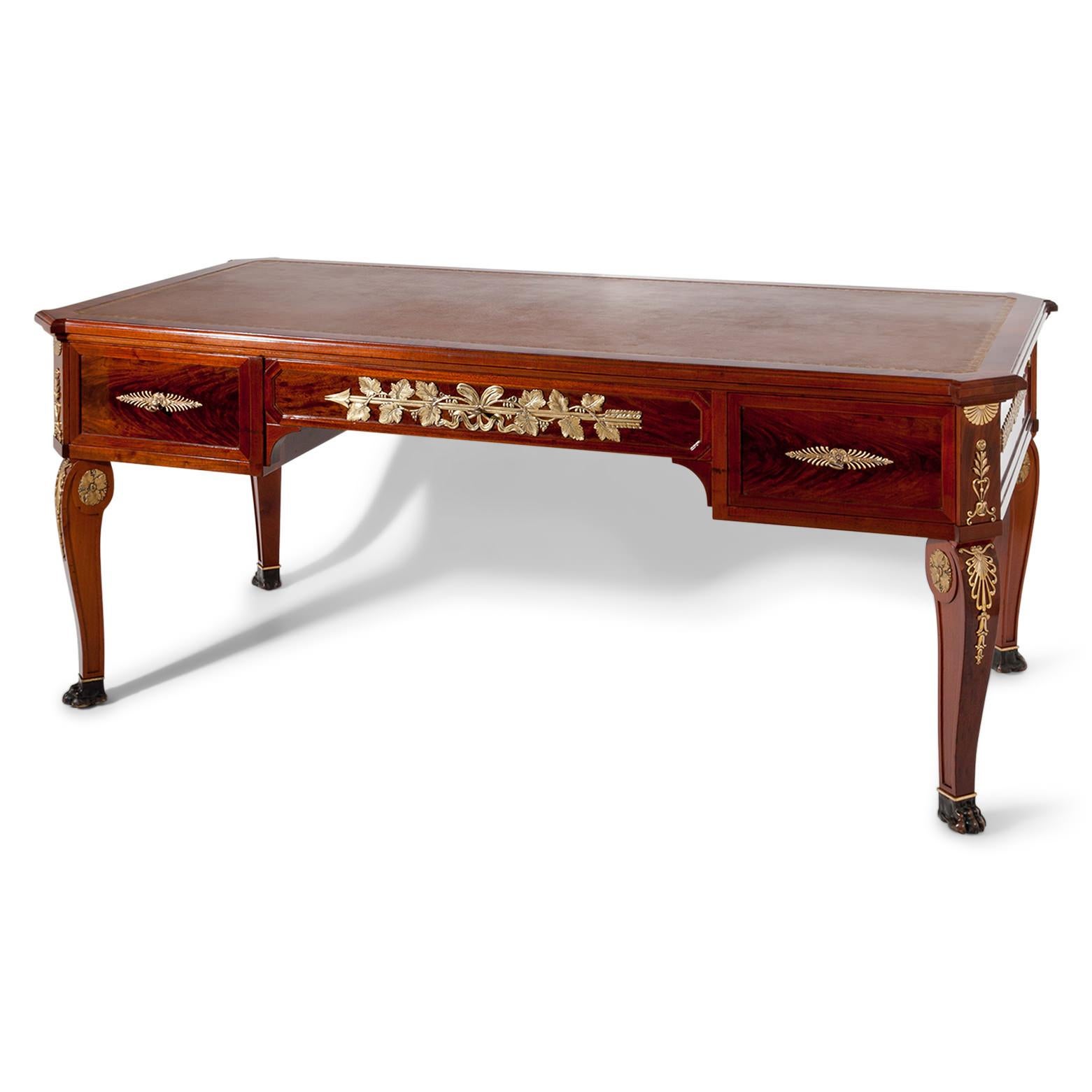 Bureau plat standing on curved legs with blackened lion’s paws. The desk has three drawers and is decorated with gilt brass fittings. The tabletop with slanted edges is covered with a brown leather.