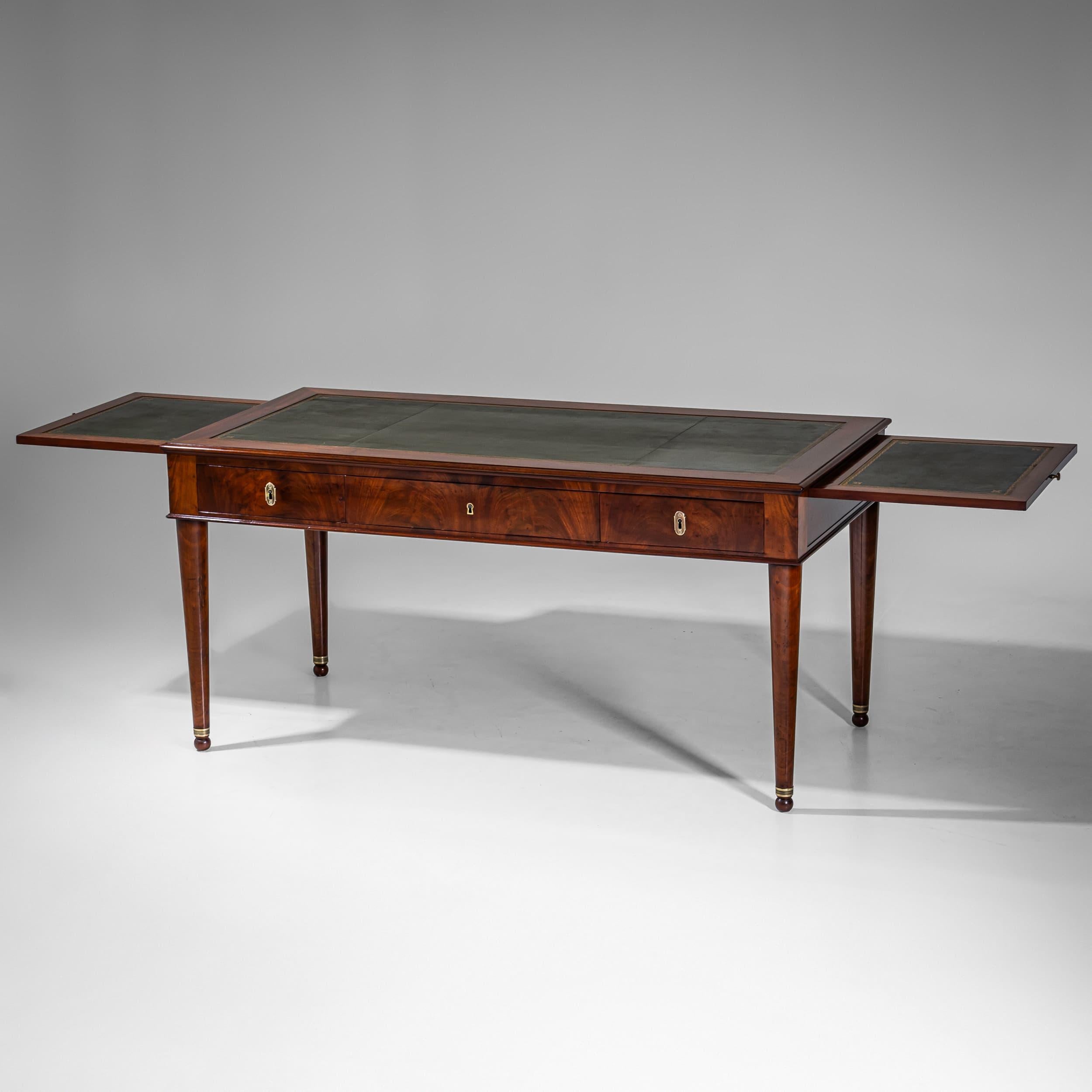Elegante scrittoio Impero (Bureau Plat) in mogano. La scrivania poggia su gambe affusolate e lisce con piedini a sfera e sabot in ottone e dispone di tre cassetti e due allunghi laterali estraibili (lunghezza massima: 260 cm). La superficie di