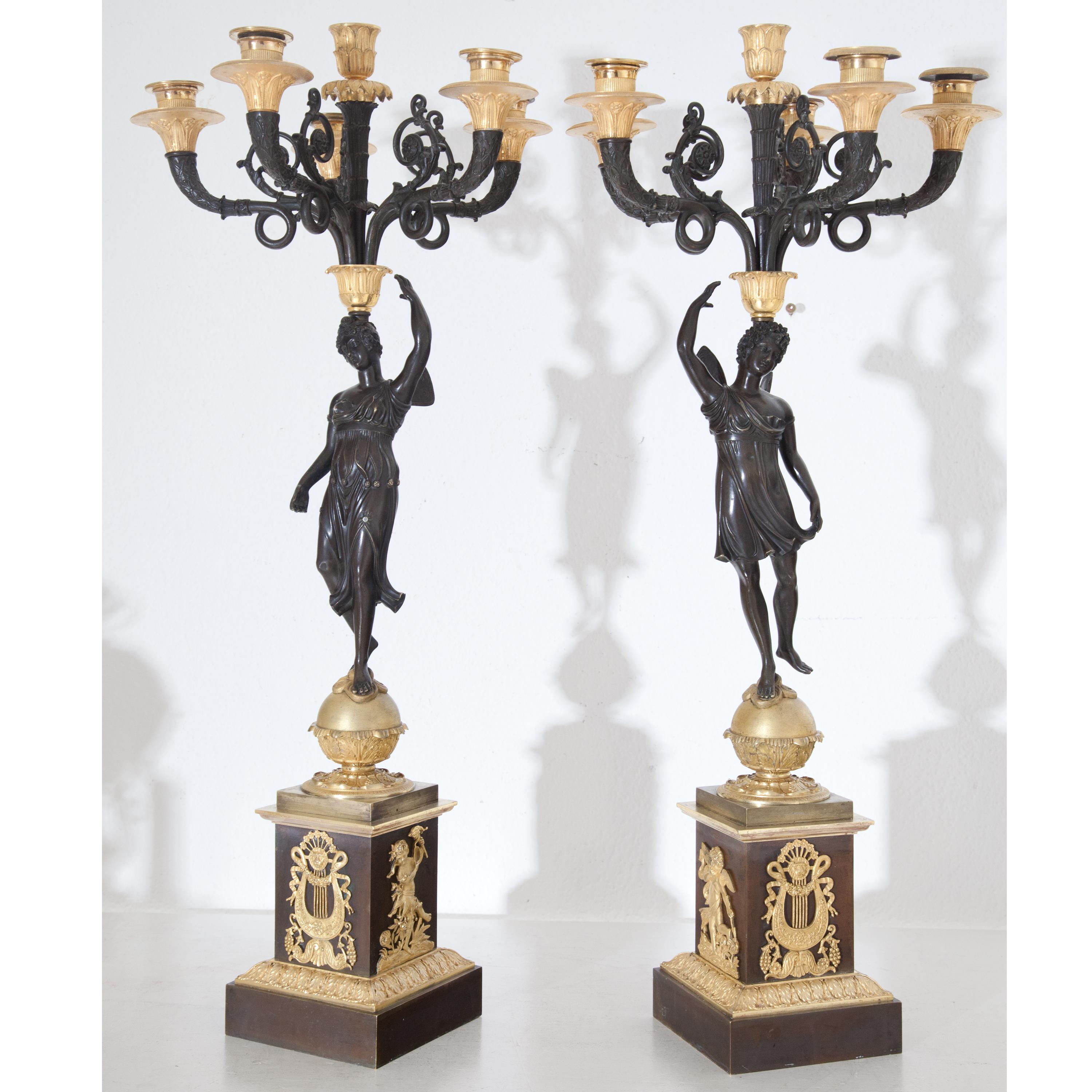 Pair of six-flamed girandoles with winged psyches on spheres, balancing the chandelier on their heads. Burnished square pedestals with lyre and Amoretti decoration. Bronze, fire-gilt. (measurements plinth: 13 x 13 cm).