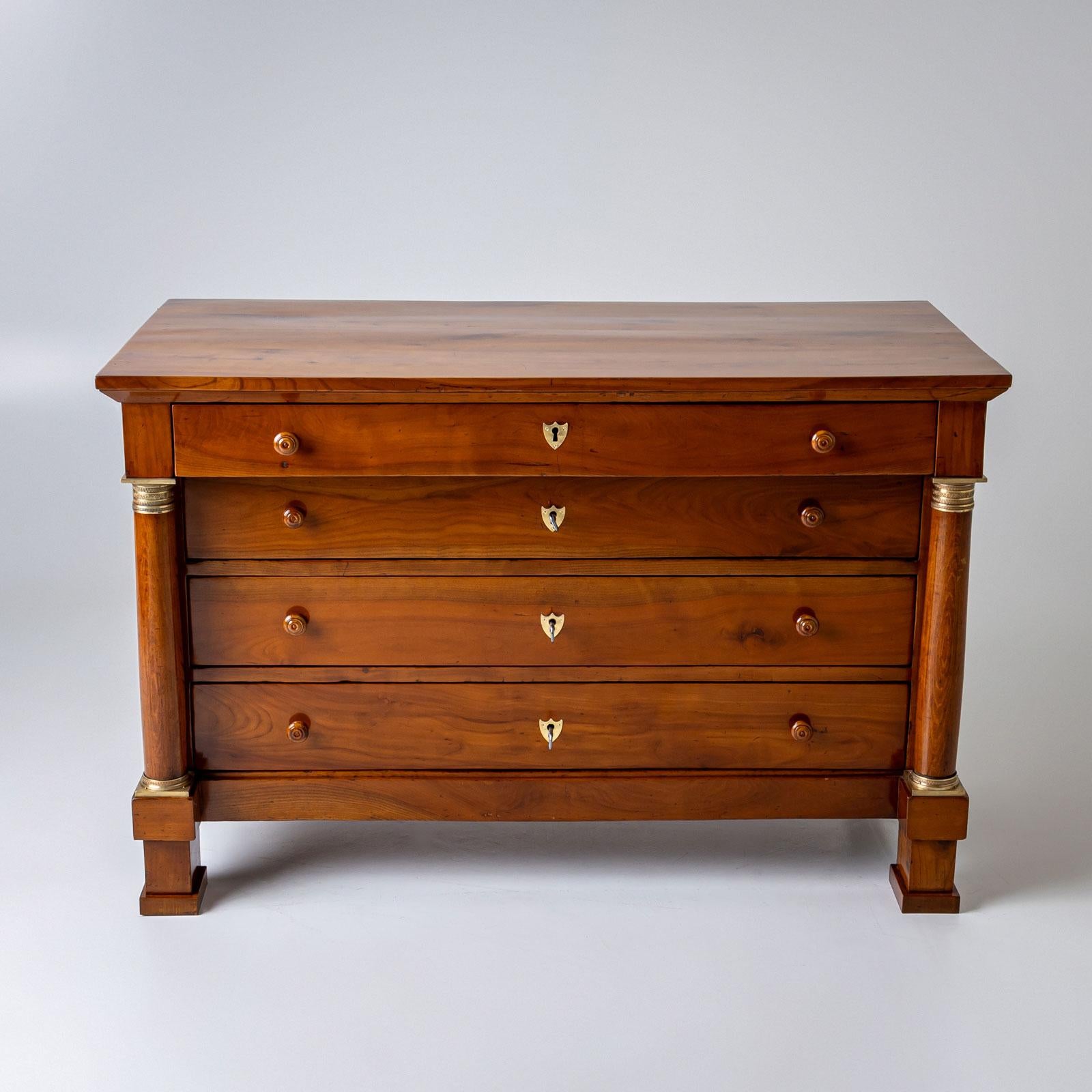 Grande cassettiera in legno di ciliegio con quattro cassetti e scudi in metallo a forma di stemma e maniglie in legno. Il cassettone è affiancato da colonne piene e lisce con capitelli e basi in bronzo. I capitelli e le basi sono decorati con
