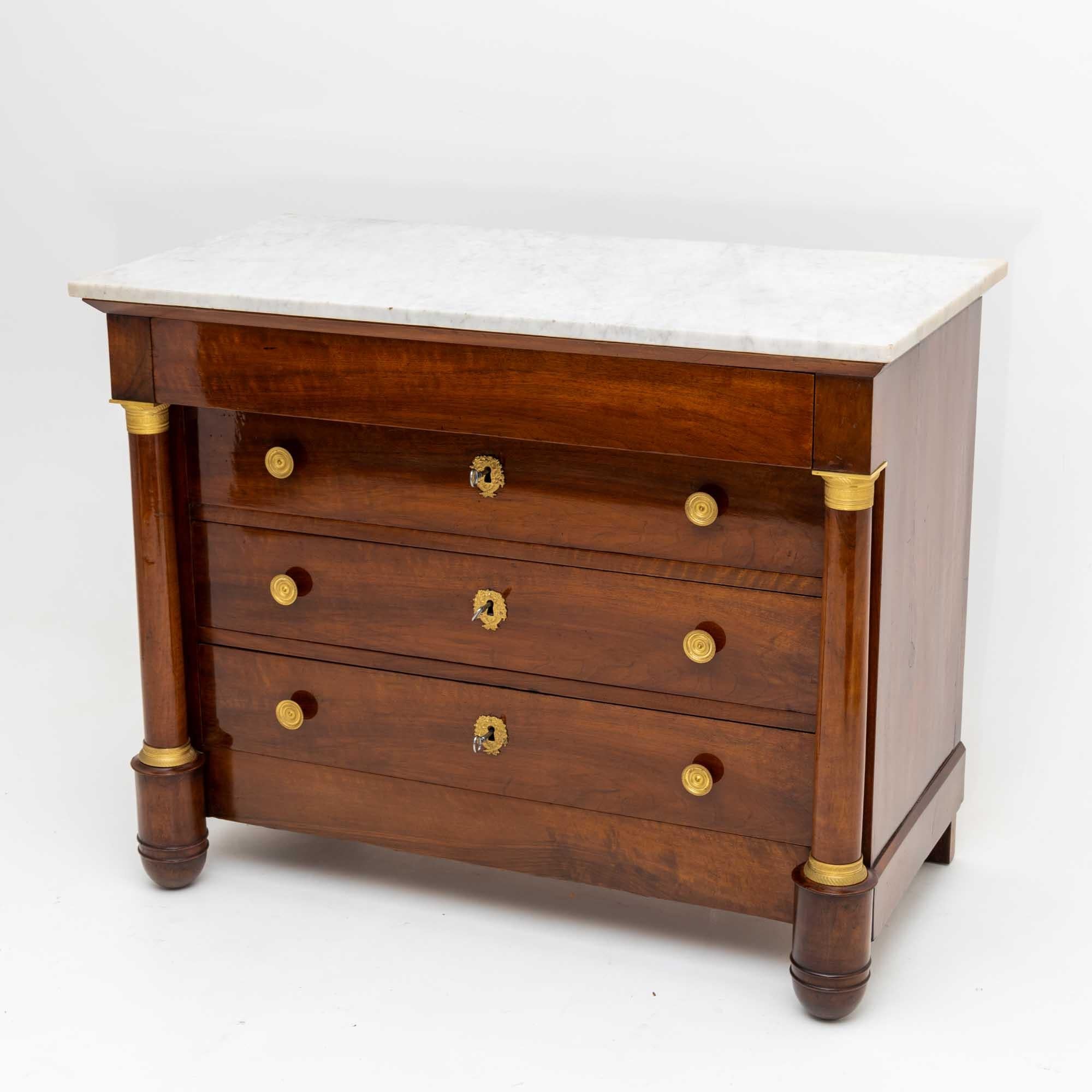 French Empire chest of drawers veneered in mahogany, with four drawers and white marble top. The corpus is flanked by columns with fire-gilded bases and capitals, which merge into the conical front feet. The head drawer rests on the capitals and has