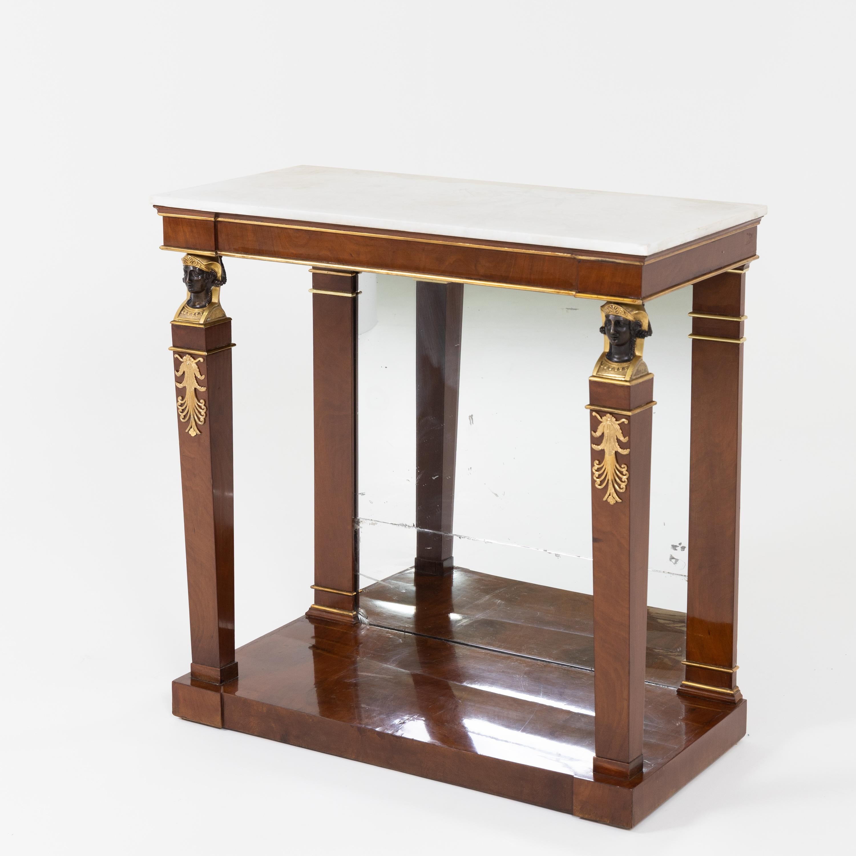 Empire console table on rectangular pedestal with partially gilded, blackened caryatid heads at the front and pilasters with brass mouldings at the back. The back wall has an old mirror glass. The white marble top rests on a straight frame with