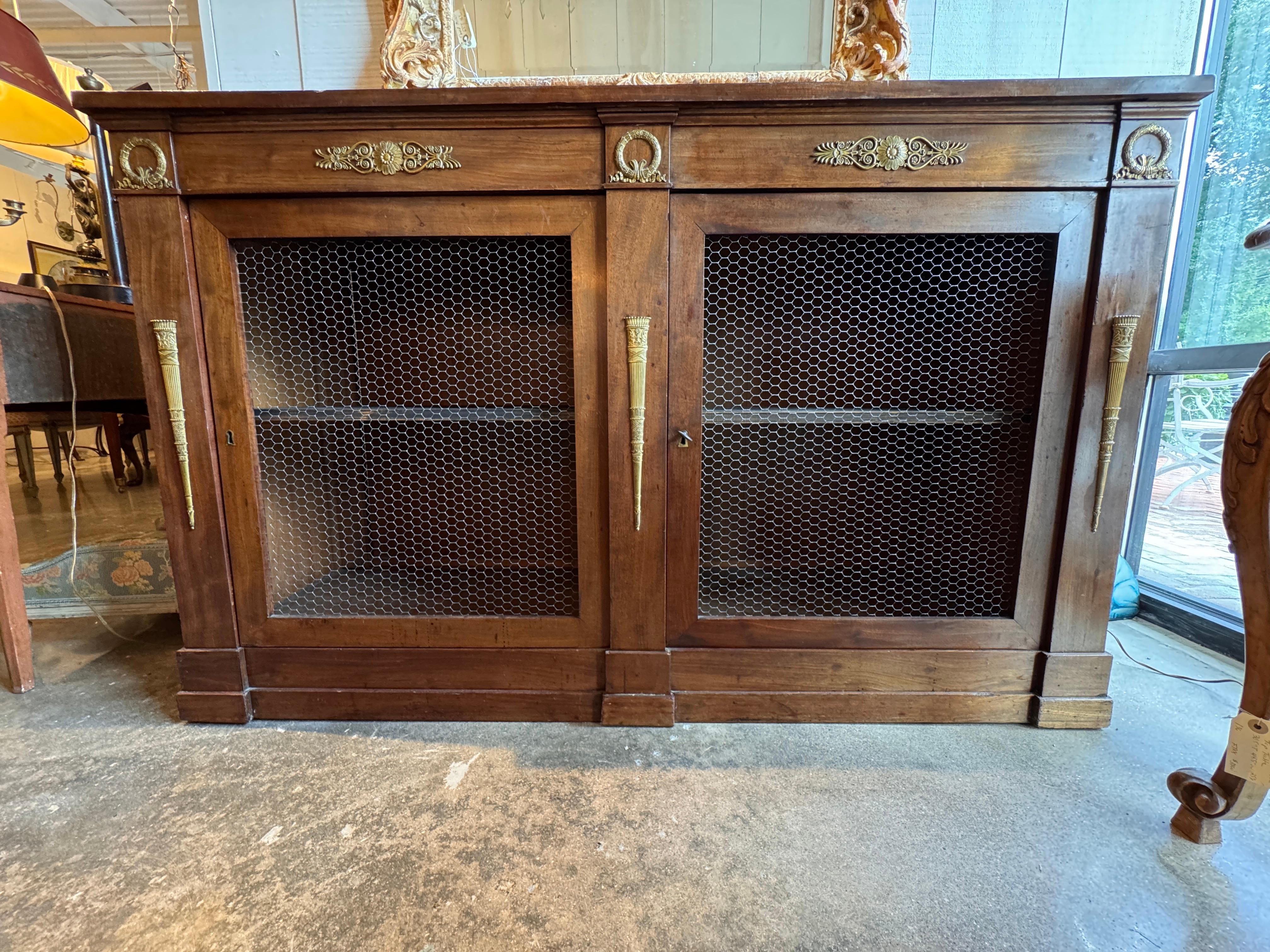 Credenza Impero con due porte anteriori in rete metallica, incorniciate da decorazioni in bronzo dorato a forma di ghirlande, rosette e torce affusolate. Il piano è dotato di un unico cassetto a fregio e l'interno presenta un ripiano centrale.