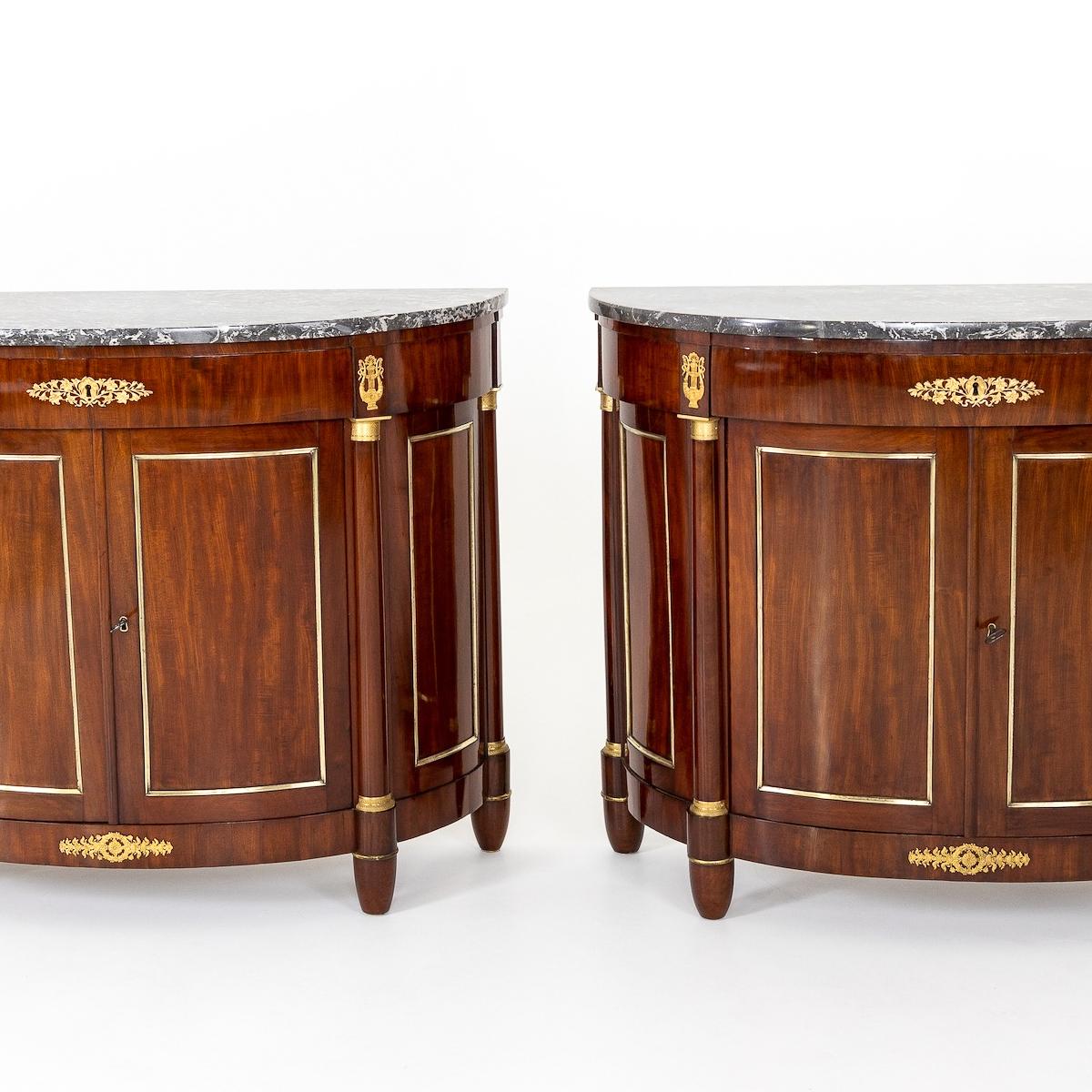 Two-door demi lune sideboards on conical feet with grey and white marble tops and bronze fittings in the form of lyre and floral motifs. The panels are framed with brass mouldings and are articulated by half-columns with bronze bases and capitals.