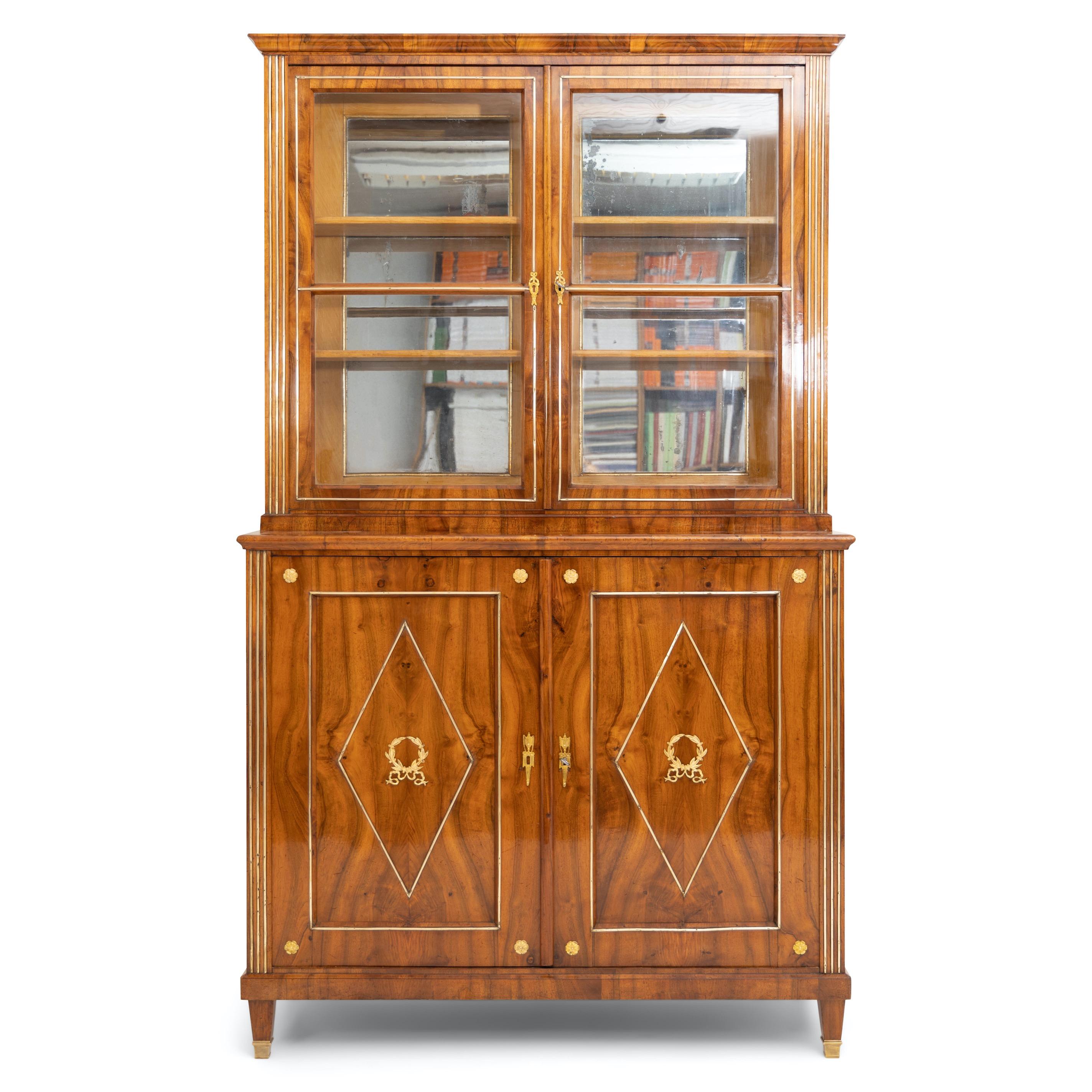 French Empire display cabinet standing on square pointed feet with brass sabots, veneered in walnut. The four-door corpus is decorated with fluted corners with brass profiles, rosettes, and wreaths with ribbon decoration on the filling panels. The