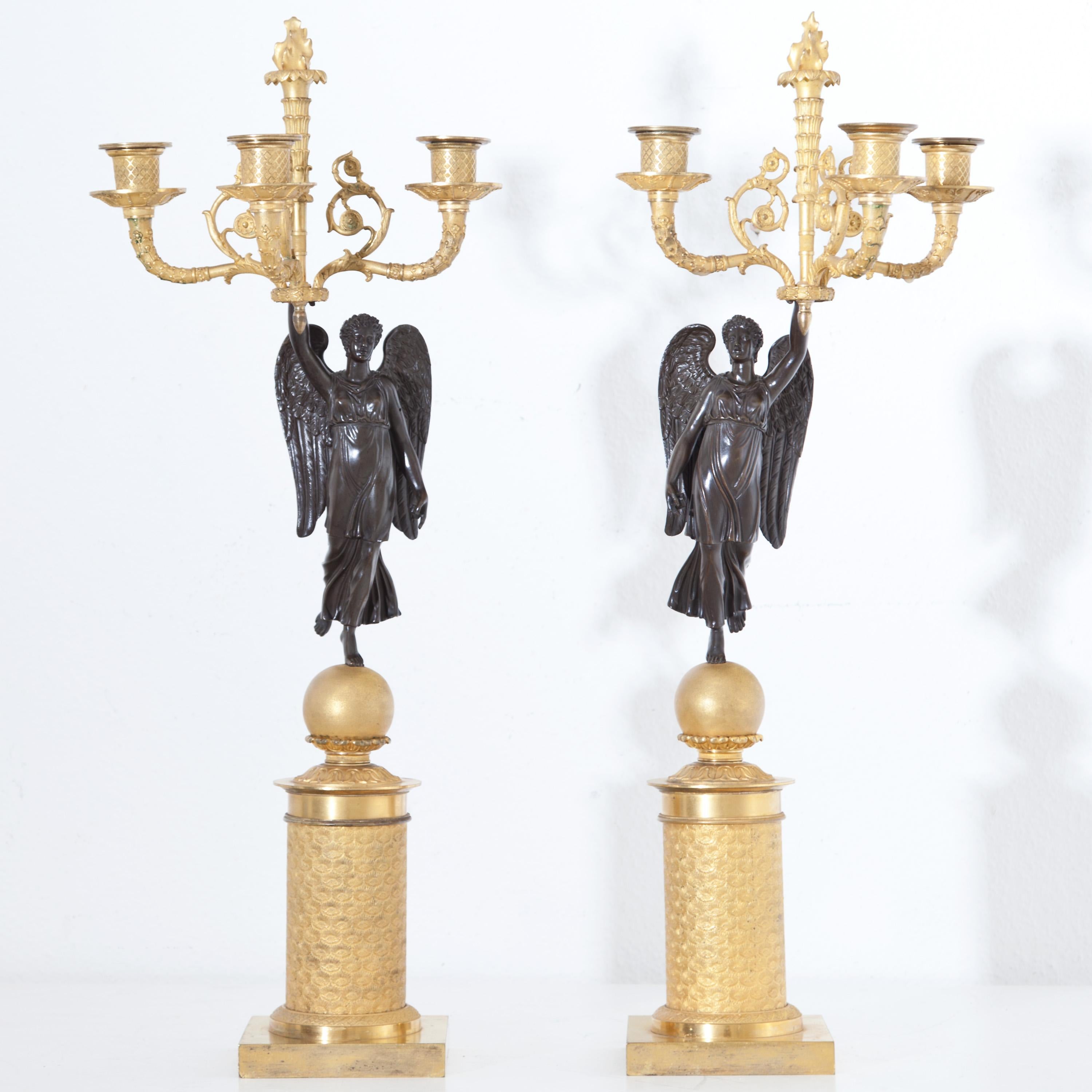 Pair of Empire Girandoles made of partly fire-gilded bronze with winged Nikes standing on spheres. The goddesses of victory are of opposite designs. They gracefully hold in their raised left and right hands, respectively, a laurel wreath which is