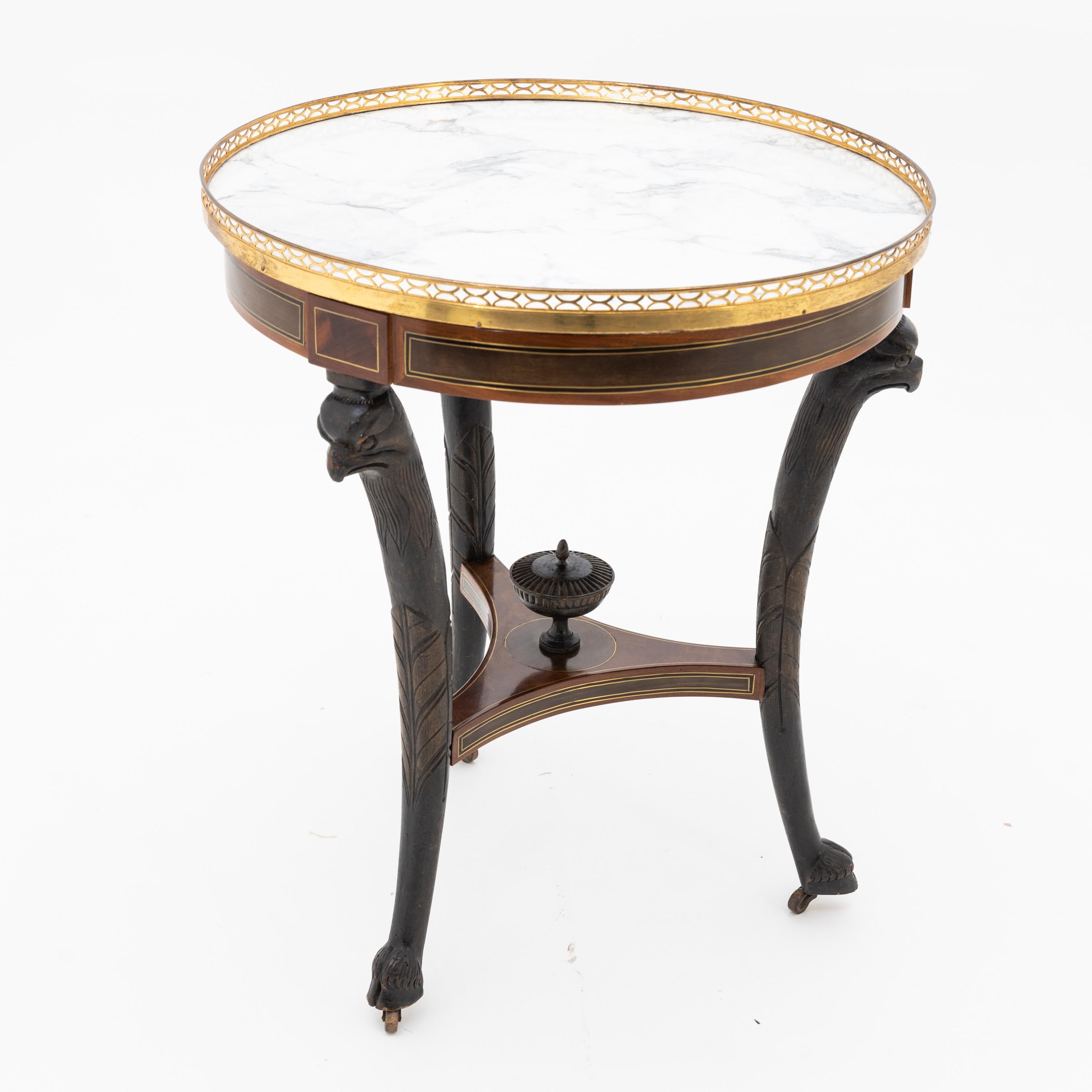 Three-legged table on casters with carved eagle heads and hooves as well as amphora decoration on the trefoil strutting. This, like the frame, is inlaid with brass threads. A low gallery surrounds the white marble tabletop. Mahogany veneered,
