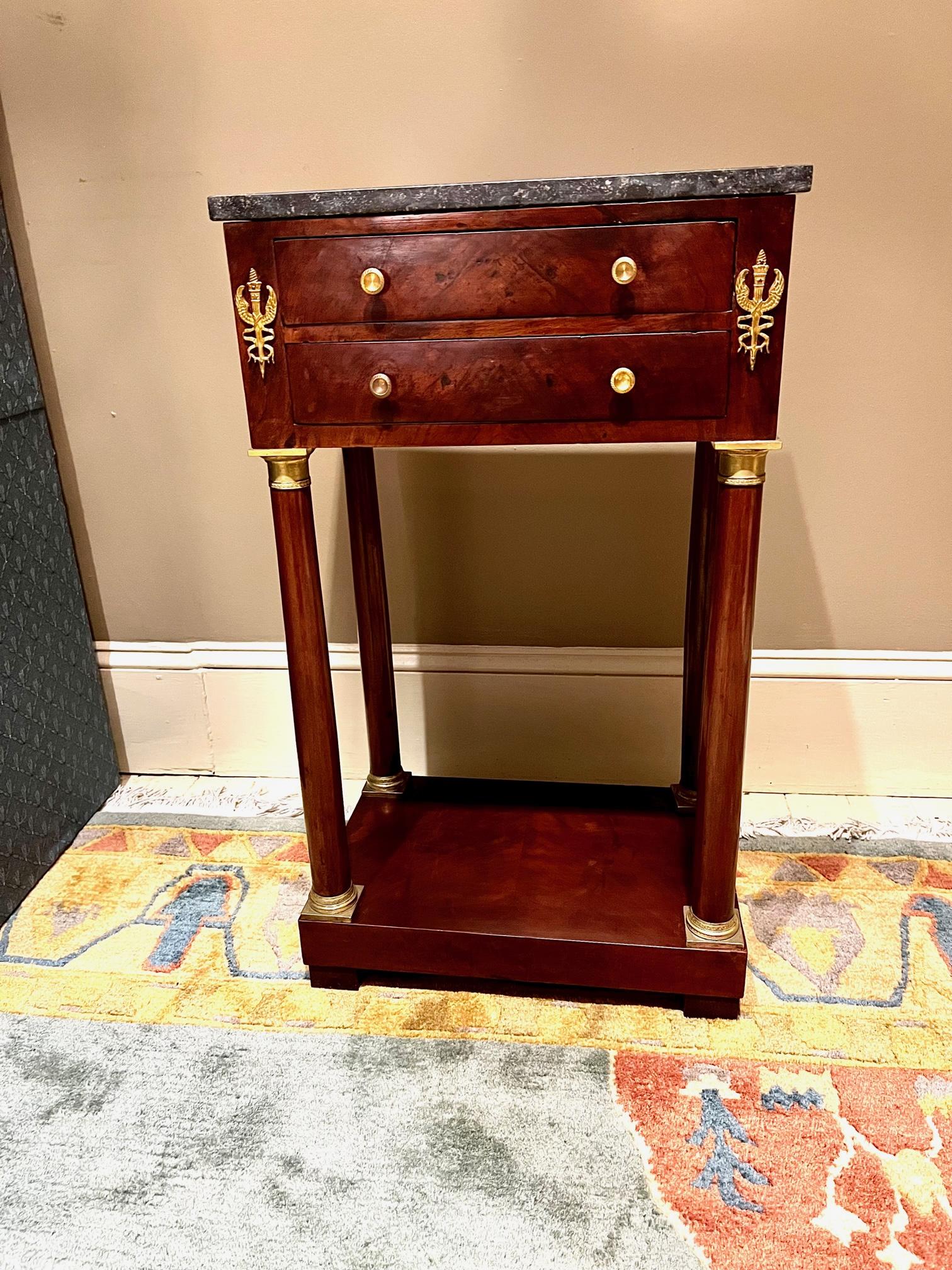 This high style 2-drawer stand is made of mahogany and mahogany book matched veneer with French pine secondary wood. The piece has a finished back, so it can be used away from the wall.  It has hand-cast gilt brass (ormolu) mounts and pulls. The top