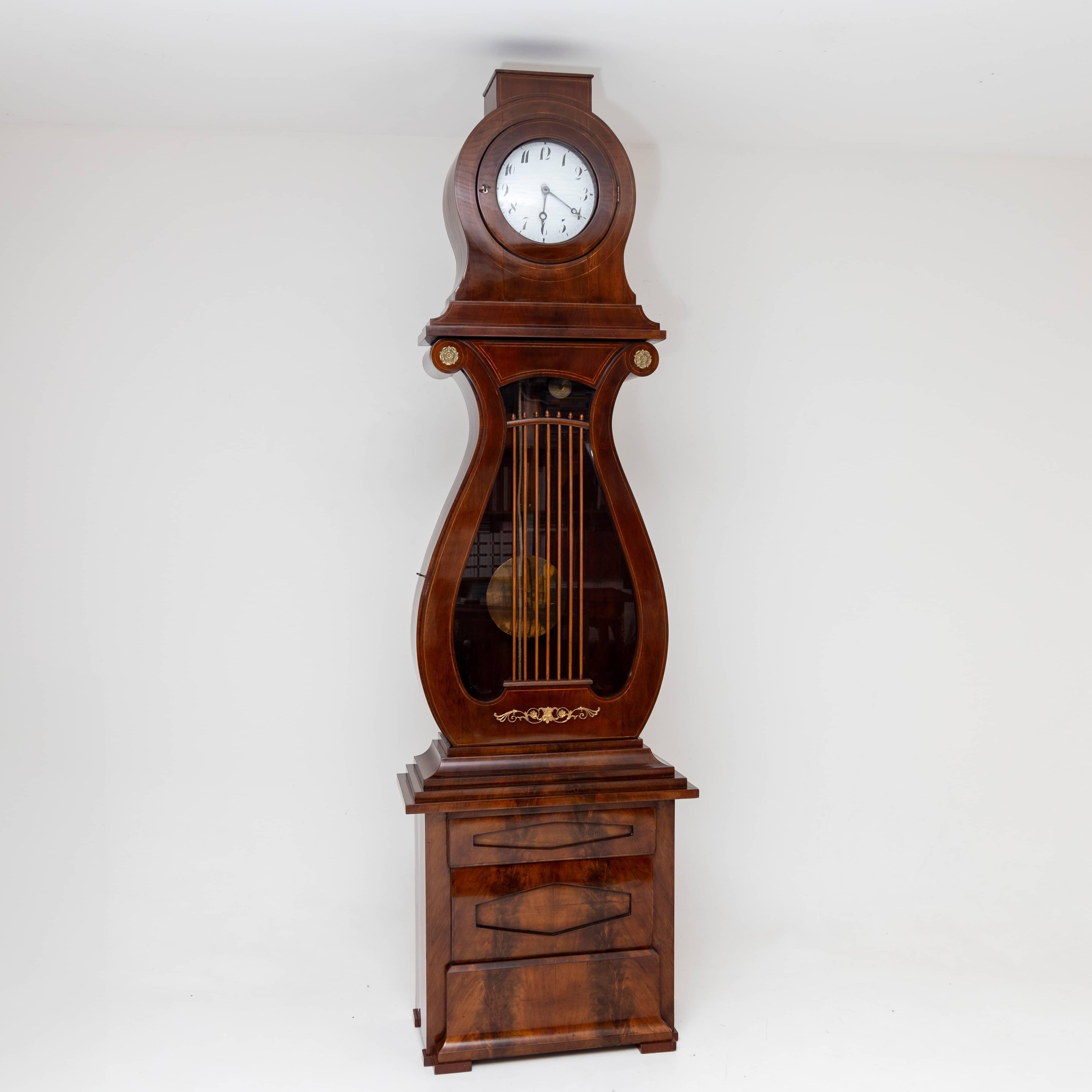 Large Empire grandfather clock with a mahogany-veneered case in the shape of a lyre. The grandfather clock stands on a chest with three drawers and diamond-shaped fillings. Above it rises the lyre-shaped pendulum case with strings out of wooden bars