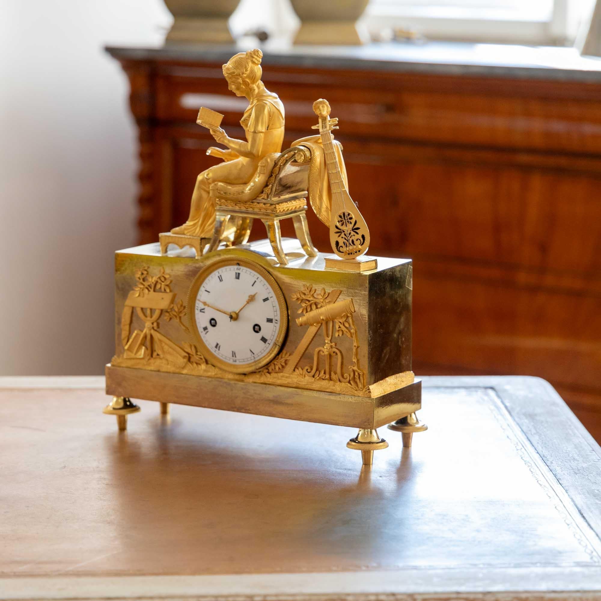 Empire mantel clock with enameled dial and fire-gilt case. The clock is crowned by a full-figure young lady sitting on a Klismos chair and reading a book. Her feet rest on a stool and a lute leans against her chair.