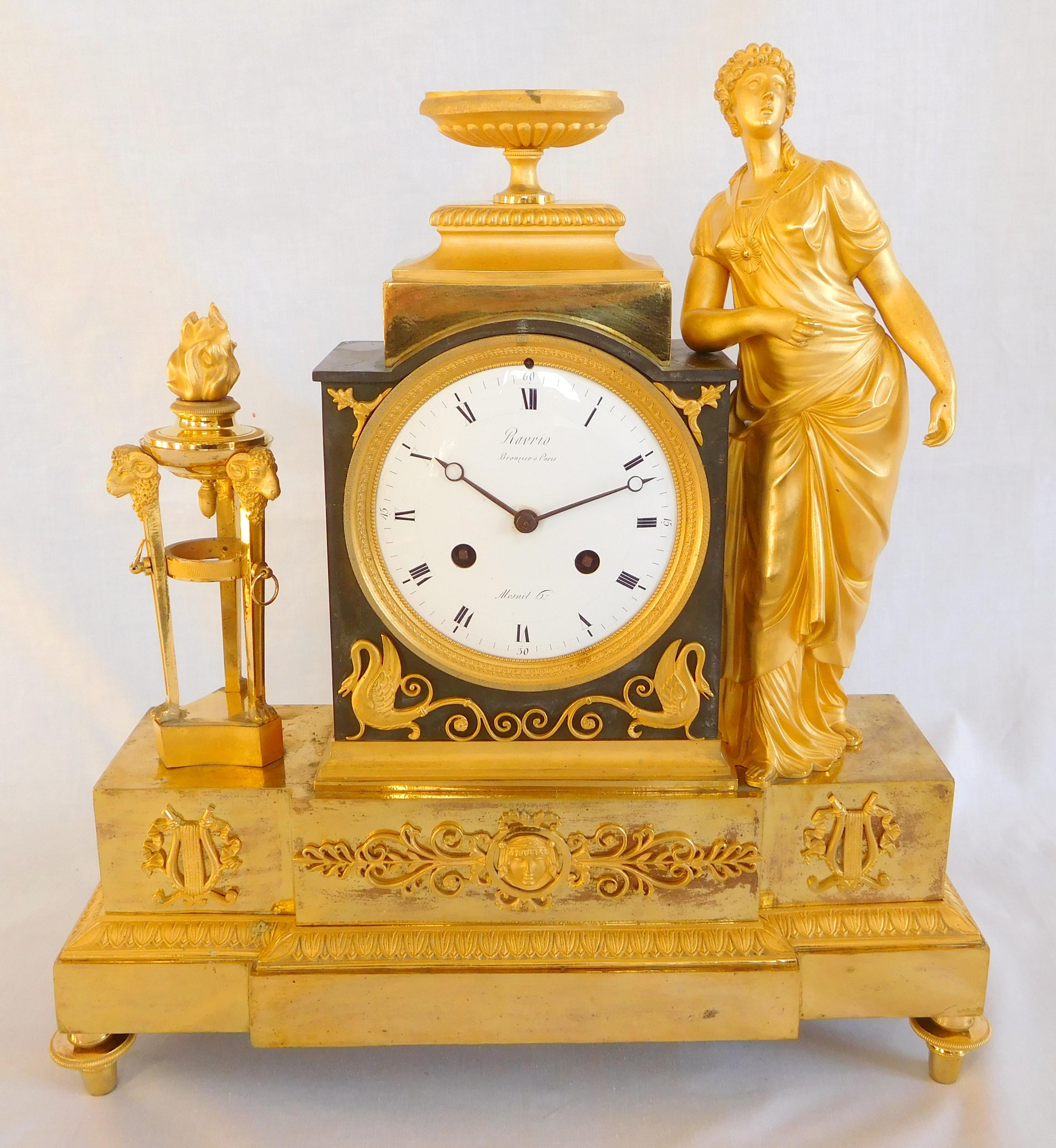 Reloj de chimenea de bronce patinado y bronce dorado al mercurio en el que aparece una virgen vestal vestida a la antigua usanza apoyando los codos en el altar del Amor. La esfera esmaltada está firmada por Ravrio (broncista) y Mesnil (relojero).