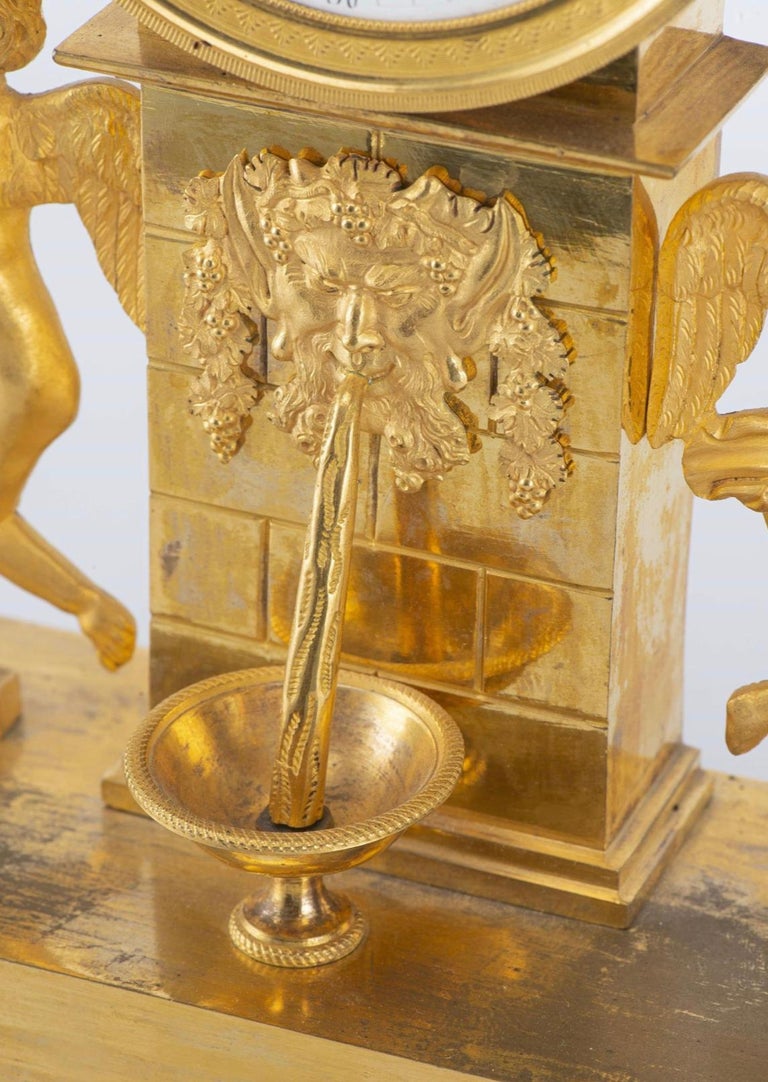 Empire period gilt bronze mantel clock decorated with putti, fountain column base and pediment with torch, swans and sprays of flowers. 

White enamelled clock face with Roman numerals for the hours and Arabic numerals for the quarter hours.