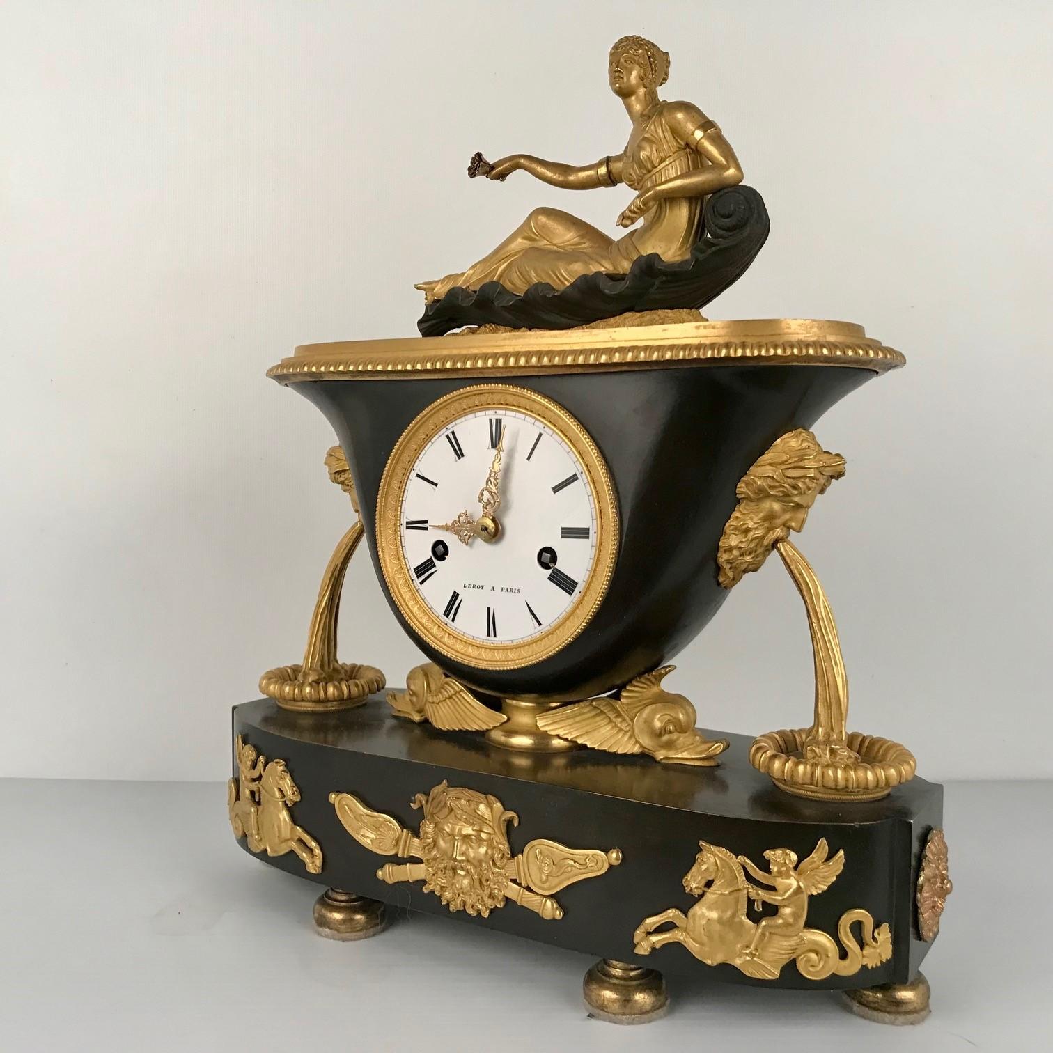 This mantle clock is by a well-respected Paris maker in a fashionable style. The oval base supports a vase shaped clock case in turn supported by winged dolphin heads and flanked by stylised fountains. The top of the clock is modeled as a young