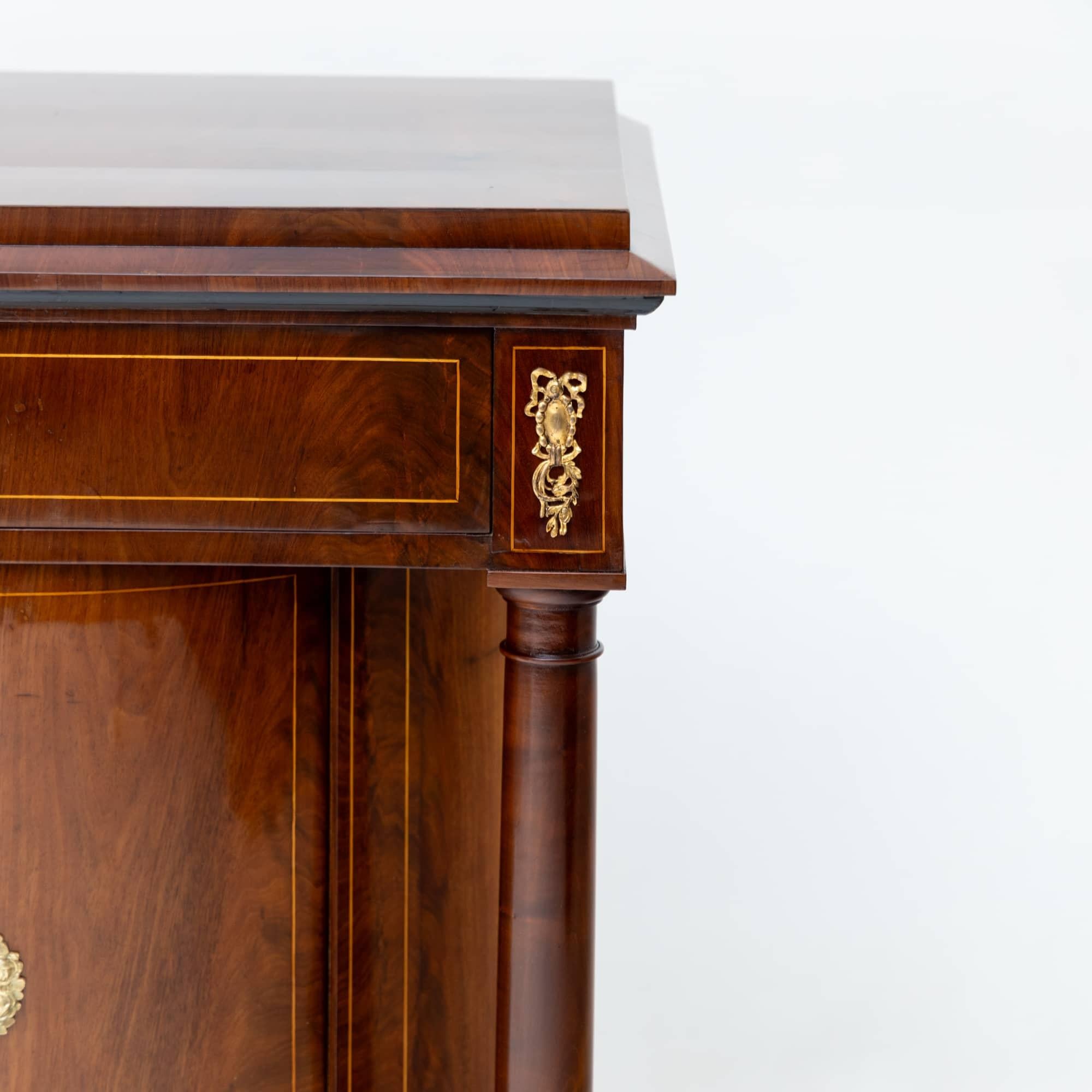 Mahogany veneered half cupboard with fine thread inlays and bronze applications. The half cupboard stands on low square feet and above a plinth drawer rises the semi-circular corpus with a door and flanking solid columns. The top is formed by