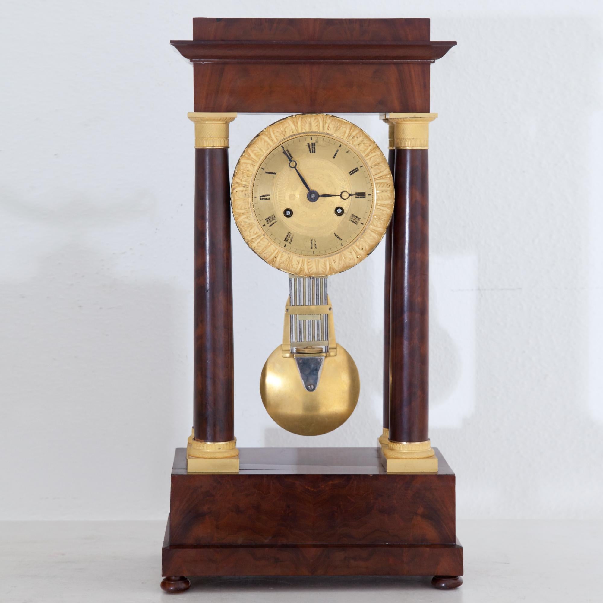 Portal clock on a rectangular, stepped base in mahogany veneered with small bun feet. The stepped architrave is supported by four smooth columns, the capitals and bases are in fire-gilded bronze. The clockwork is fixed in between the columns. Clock