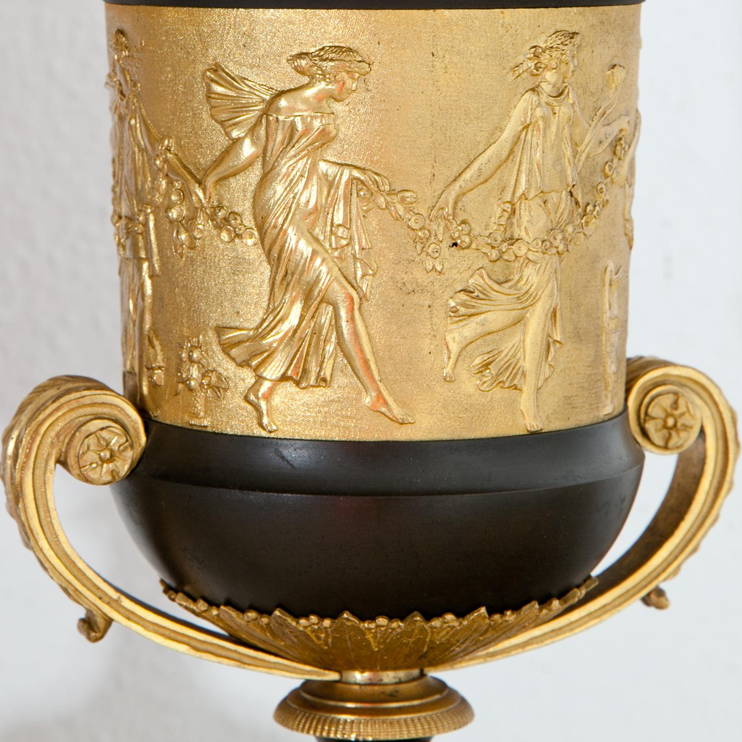 Paar teilweise vergoldete Brûle-Parfum-Urnen, Claude Gallé zugeschrieben, auf grauen Marmorsockeln mit Hermes-Maskaronen. Die Urnen sind mit einer vergoldeten Reliefwand verziert, auf der tanzende Mänaden dargestellt sind. Der Deckel und der Rand