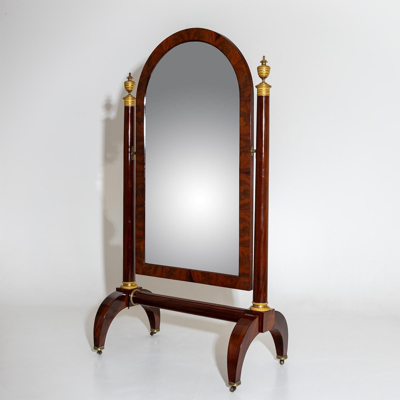 Large standing mirror with rotating mirror in an arched frame. The mirror is held by lateral columns which end in fire-gilded bronze urns. The bases are also fire-gilt. The psyche stands on forked legs with brass castors. The mirror is veneered in