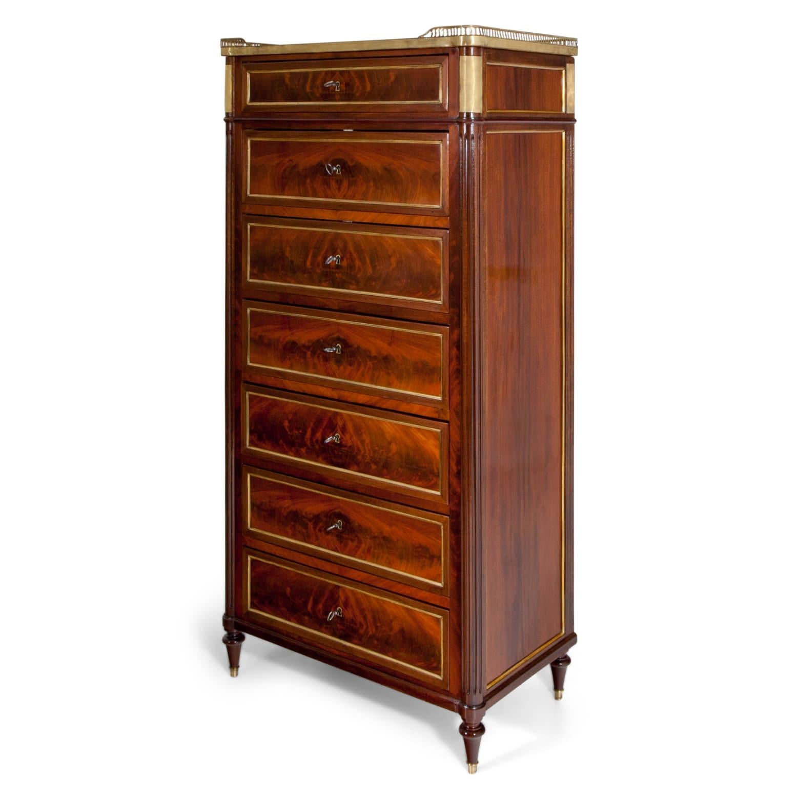 Tall chest of drawers with seven drawers and rounded, fluted corners, standing on conical feet with brass sabots. The top drawer is accentuated with brass fittings and the top is framed on three sides with a short balustrade. Each drawer is framed