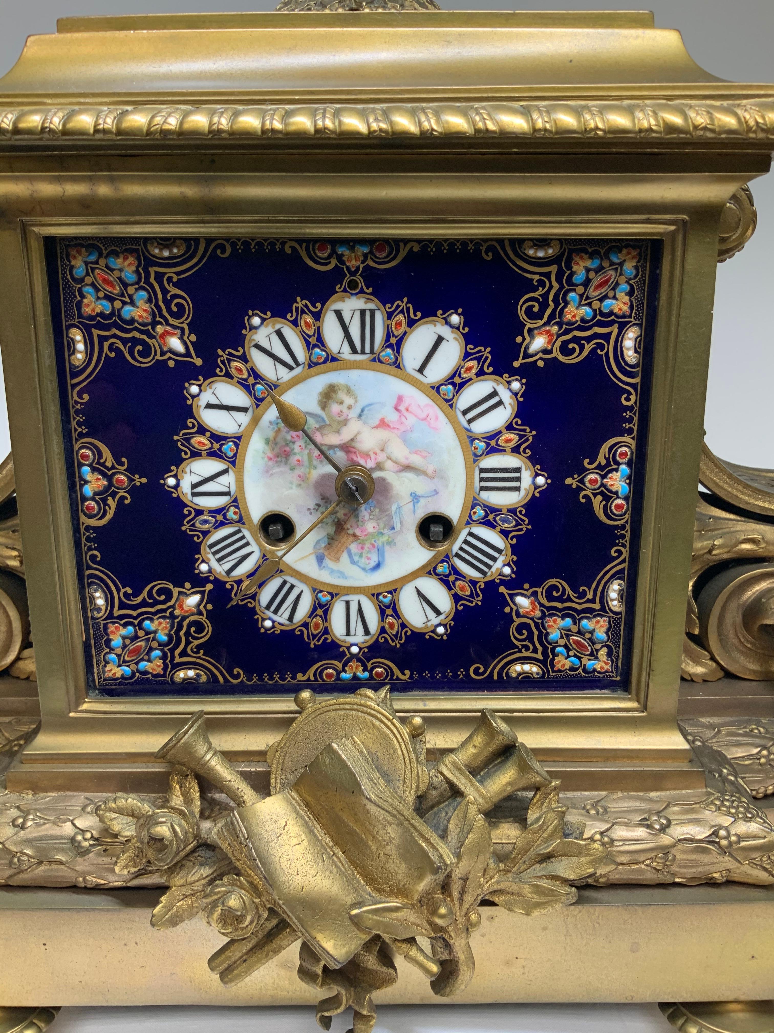 This a Sevres style porcelain ormolu bronze mounted mantel clock. The frontal cobalt blue porcelain panel is hand painted with a winged cherub holding a basket of flowers. Below him, there is another basket of flowers with ribbons. The dial is in