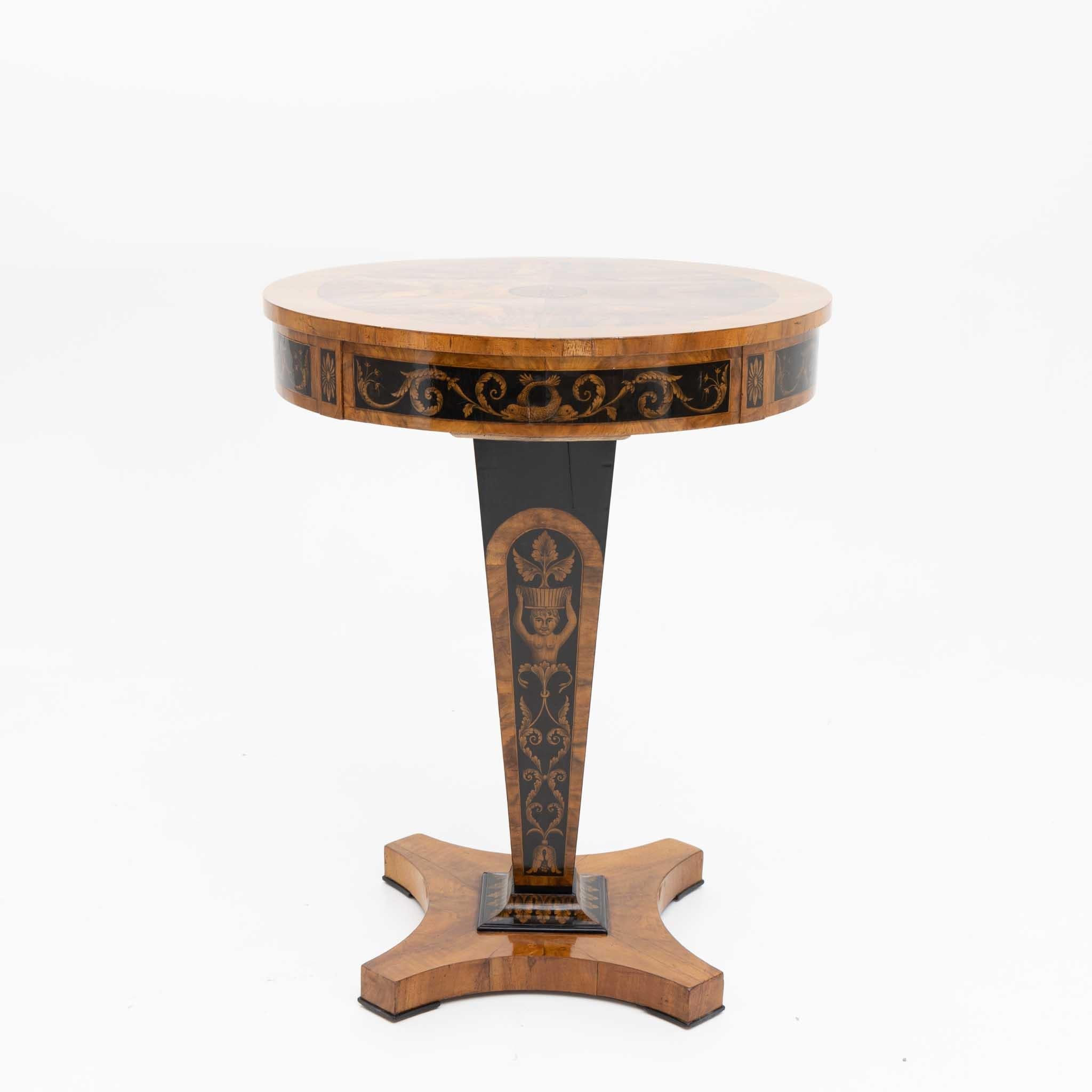 Round Empire side table with one drawer. The table is veneered in walnut and shows black ink painting with dolphin motifs and vines on the frame and on the downward tapering stand. The table rests on a quatrefoil base. It has been carefully restored