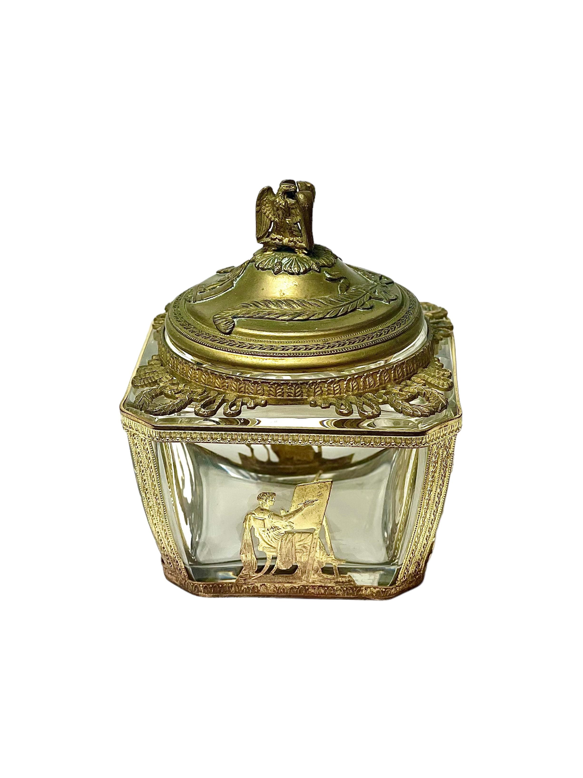 A very decorative Empire-style crystal sweet box, or Bonbonnière, intricately clad in a gilt-brass frame, with elaborately worked gilt metal mounts of the arts, a warrior on his chariot, and laurel wreaths. This square-sided crystal glass box dates