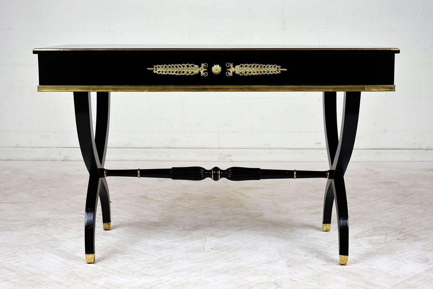 This 1950s Empire style writing desk is made of mahogany wood stained an ebonized color with a lacquered finish. There is a single drawer with brass ormolu accents that compliment the brass moulding on the edges of the desk. The stretched X-shaped