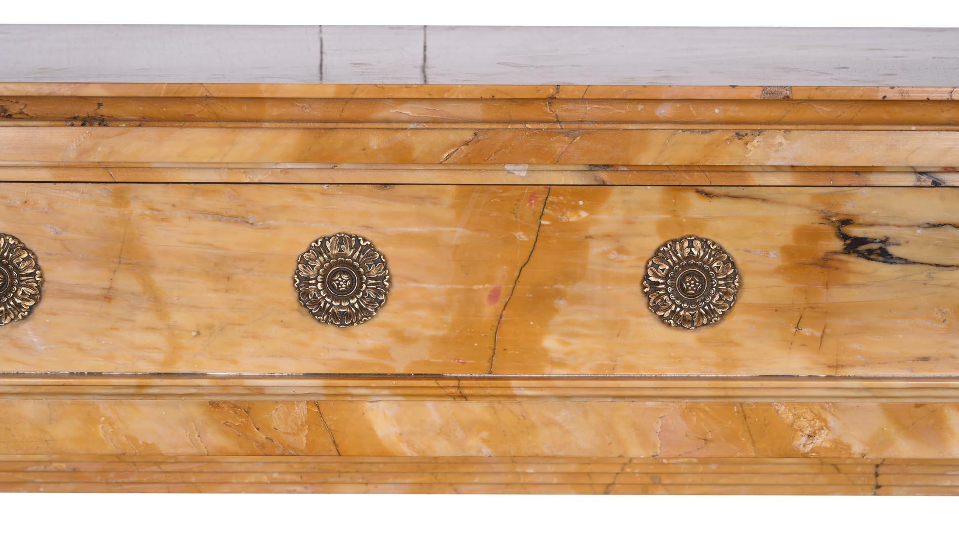 Caminetto in stile impero realizzato in marmo giallo di Siena, che si distingue per i suoi toni caldi e le venature pronunciate. Il fregio è decorato con una serie di rosette in bronzo dorato finemente cesellate. Due colonne rotonde con basi e