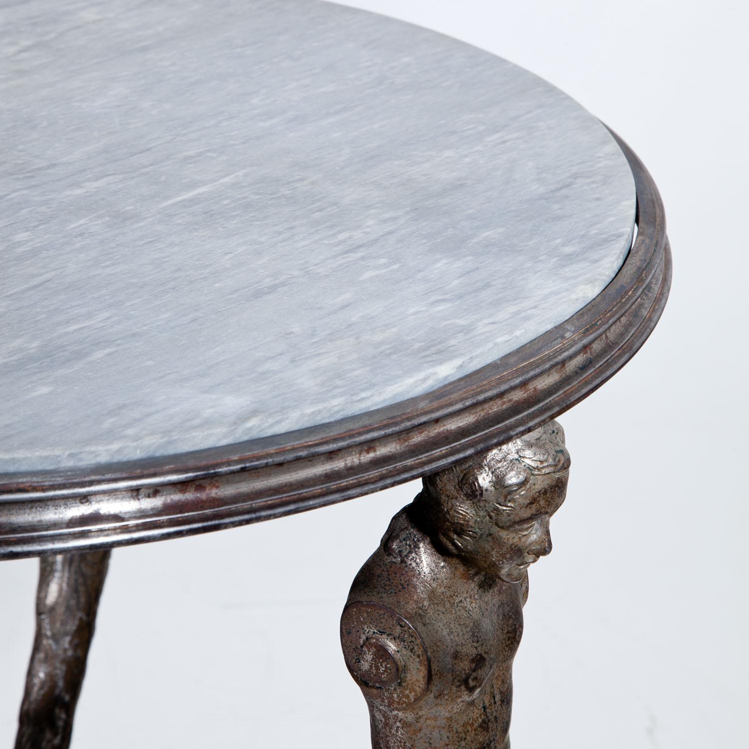 Iron table with three legs and atlantes without arms carrying the grey marble tabletop. The feet end in stylized hooves. Between the legs is a second tier with a smaller grey marble top.