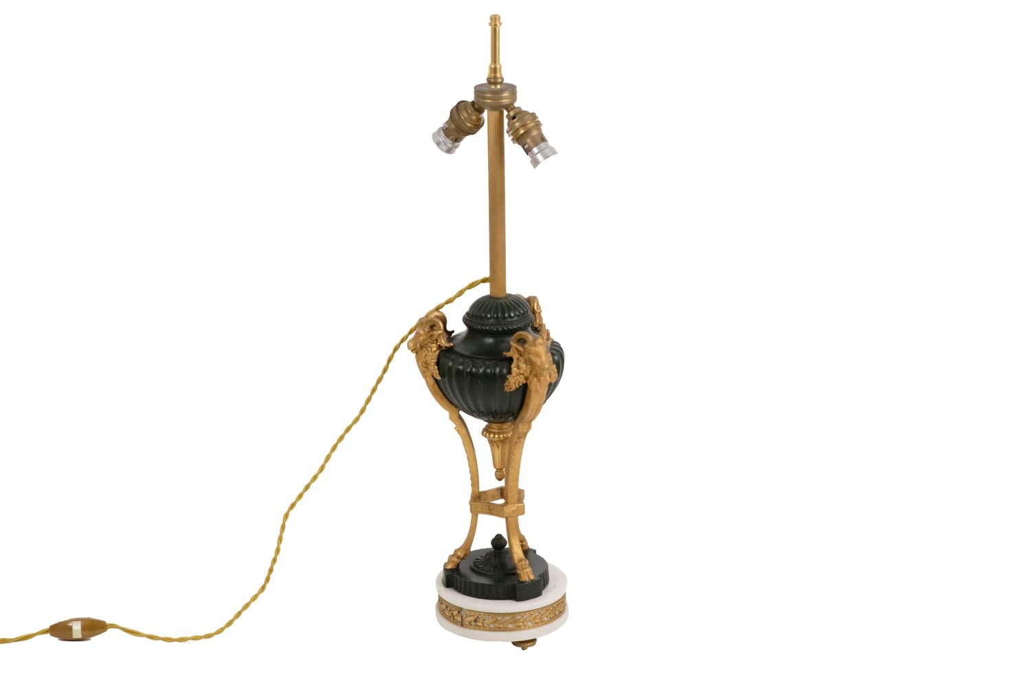 Lampe de style Empire en bronze à deux patines en forme de cassolette décorée de godrons et entourée de trois têtes de béliers terminées par des sabots. Cassolette surmontée d'une tige cannelée et terminée par une forme de tulipe décorée de feuilles