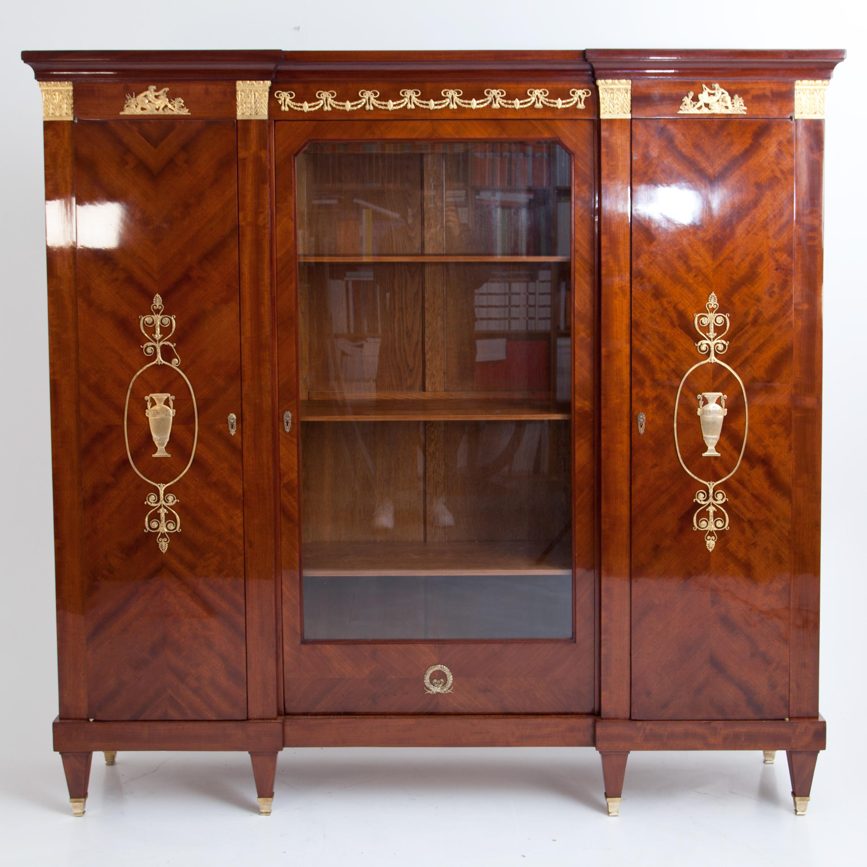 Large three-door bookcase in mahogany veneer standing on square pointed feet with brass sabots. The cupboard is decorated with large gilded appliqués in the Empire style in the form of urns, arabesques and festoons, and above the side doors are