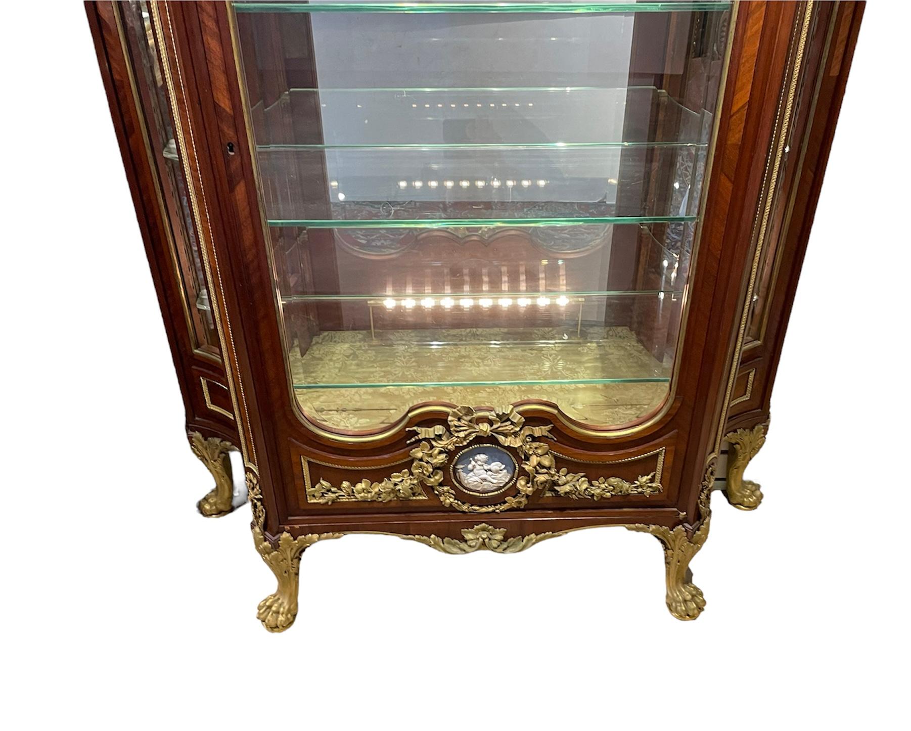 This is an Empire style gilt bronze mounted wood, glass and marble top heavy and large vitrine/curio cabinet. The wide tapered rectangular vitrine has four glass walls framed with wood. The front door is one of them and it is beveled. It is