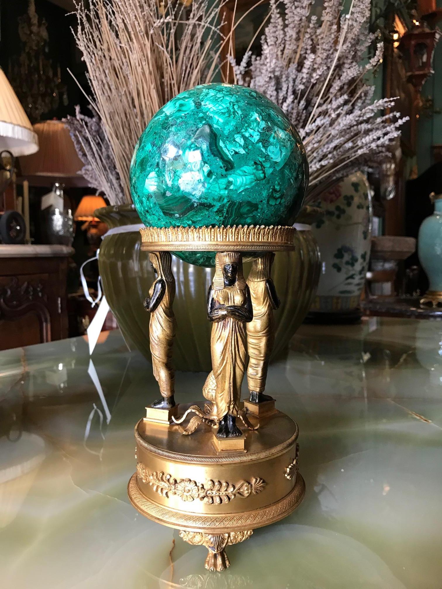 A Beautiful Empire style malachite orb on a bronze doré stand retour d'Egypte of 3 figures of standing maidens with a pine cone in the center an detailed crown holder hosting a large hand carved malachite round stone ball sphere
Malachite is a stone