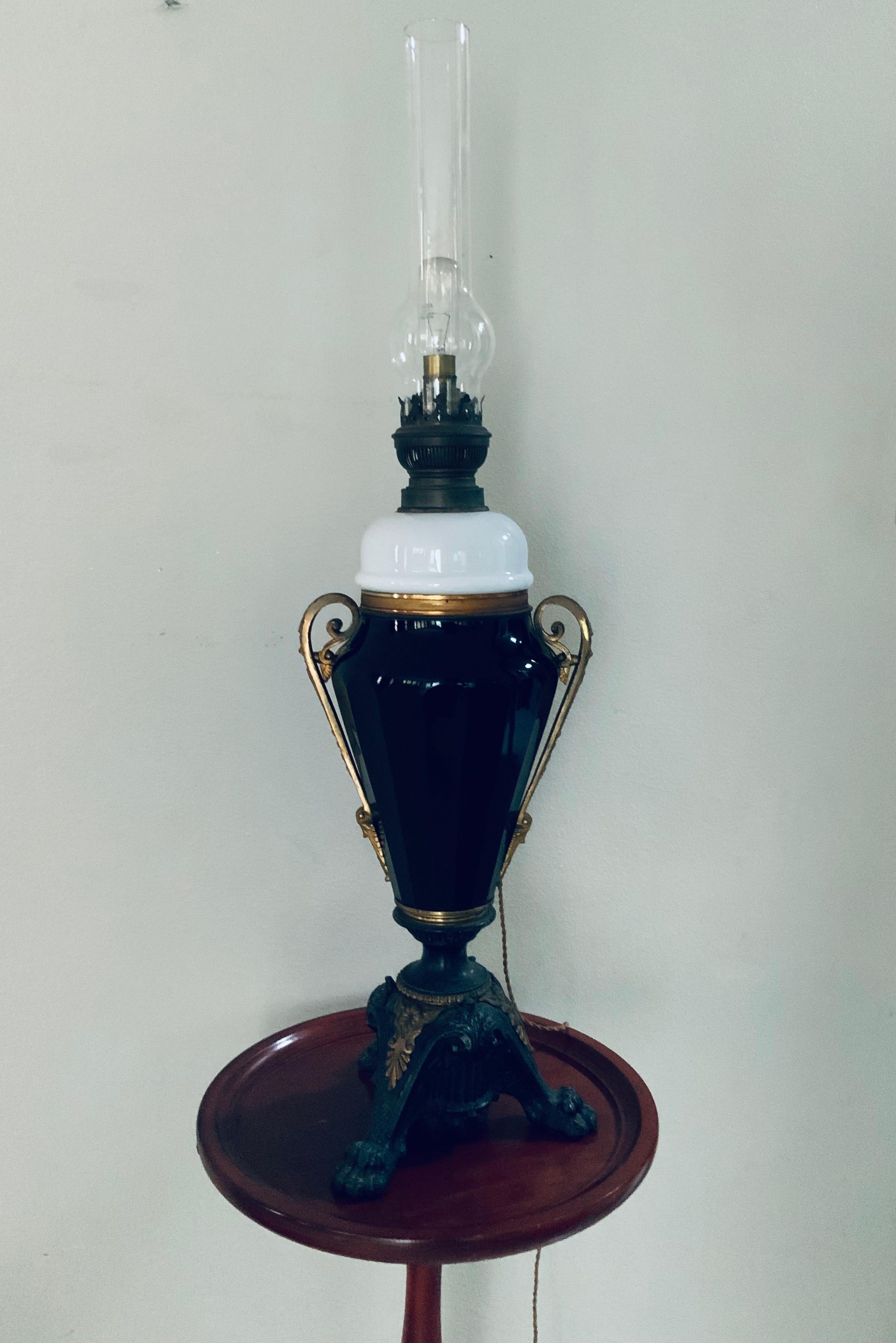 This is an Empire style tall large oil lamp that was converted to an electrical one. It consists of a cylindrical long chimney lamp clear shade attached to a brass burner holder; this in turn is connected to a semilunar shaped milk glass font joined