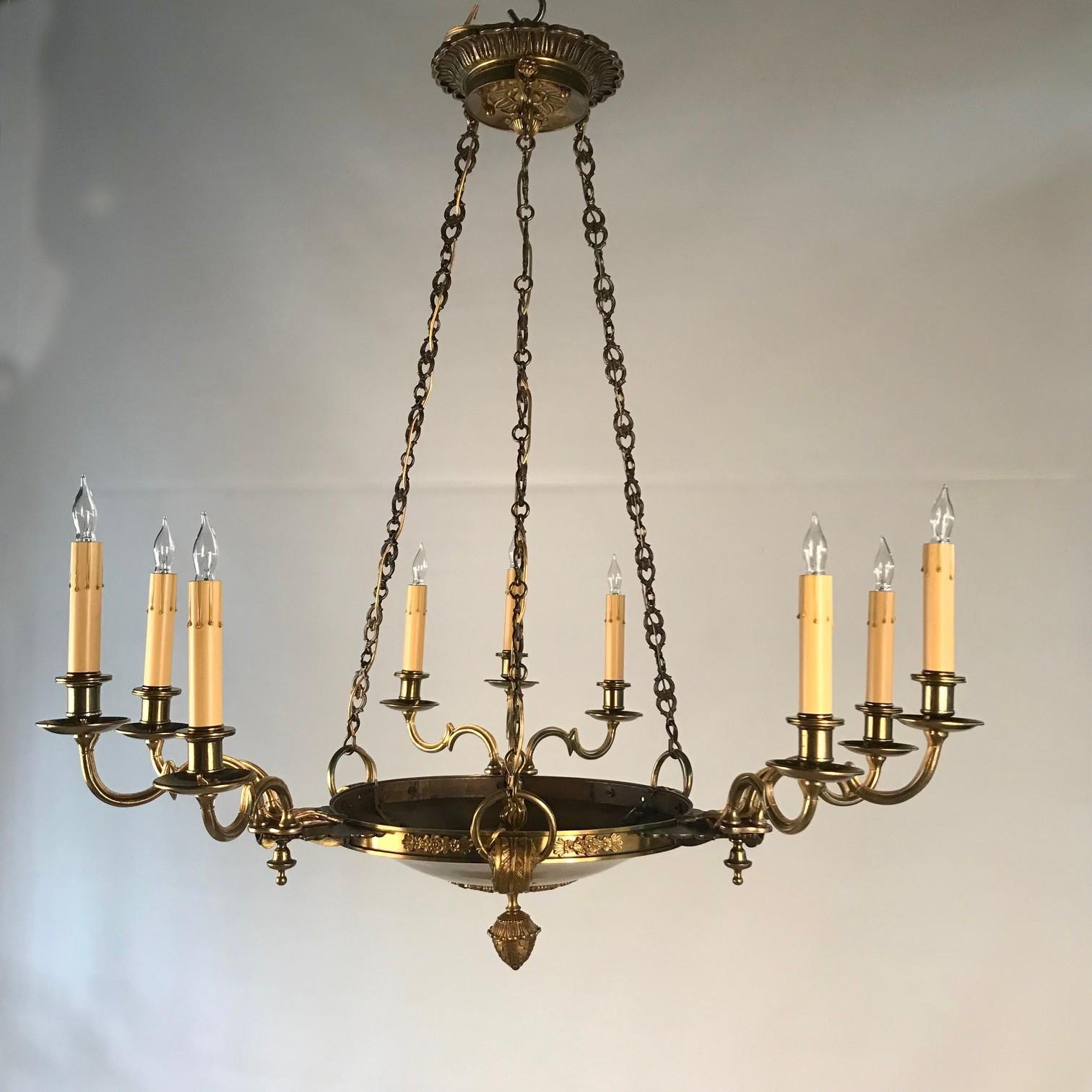 Empire style chandeliers tend to be quite hefty and large, This bronze example is unusually delicate. The dished circular pan is hung from three flat pierced suspension chains, and supports three clusters of three lights with shaped arms, candle