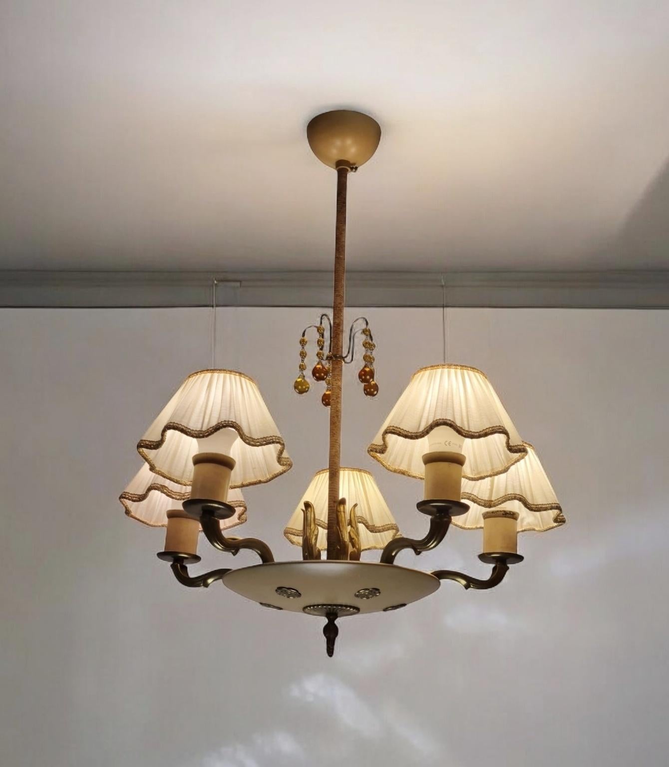 A beautiful empire style ceiling lamp in patinated brass, beige painted metal, amber glass, string decoration, and silk shades. The lamp model is 1374 by Paavo Tynell and manufactured by Taito Oy in the 1940s.

The lamp has 5 light outputs topped