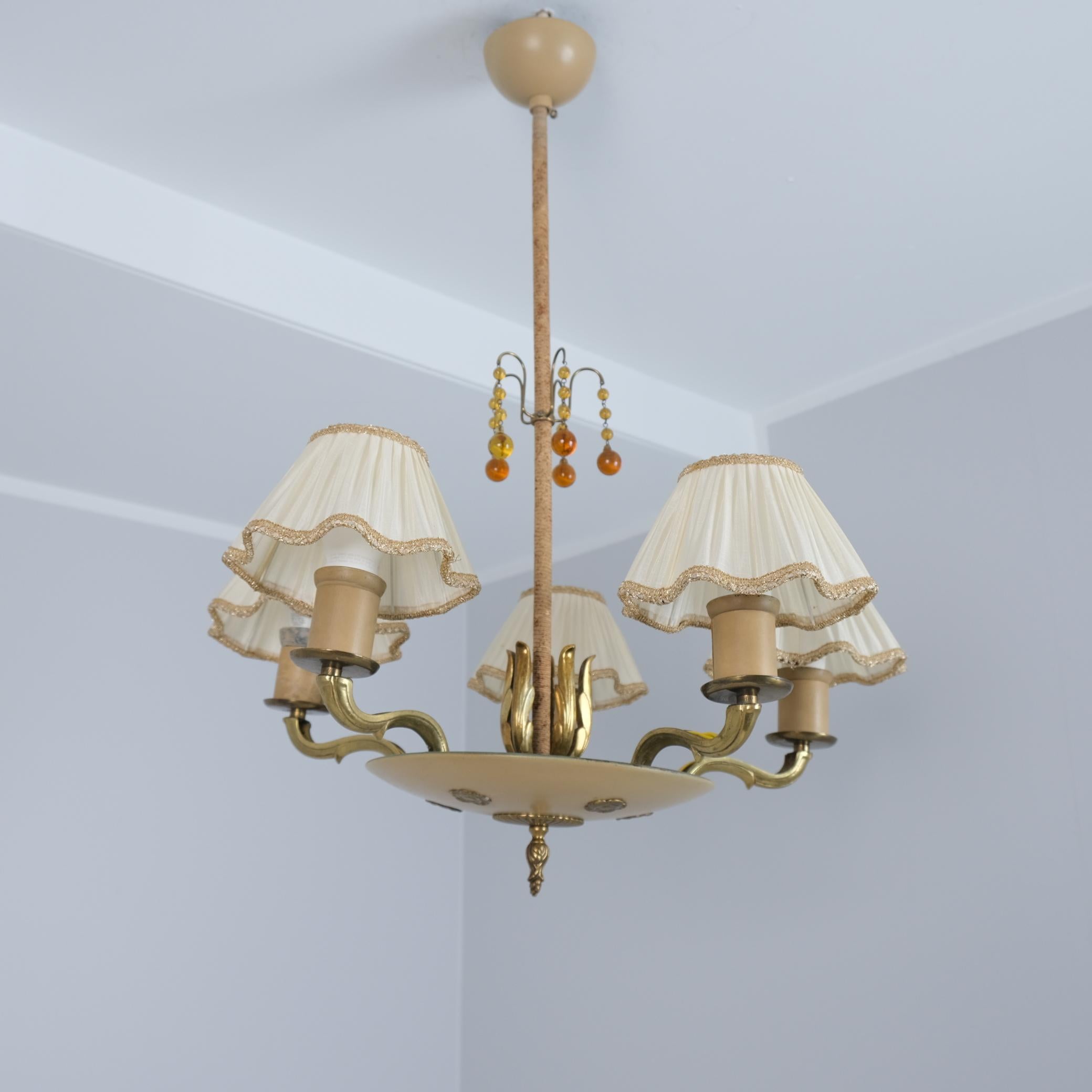 A beautiful empire style ceiling lamp in patinated brass, beige painted metal, amber glass, string decoration, and silk shades. The lamp model is 1374 by Paavo Tynell and manufactured by Taito Oy in the 1940s.

The lamp has 5 light outputs topped