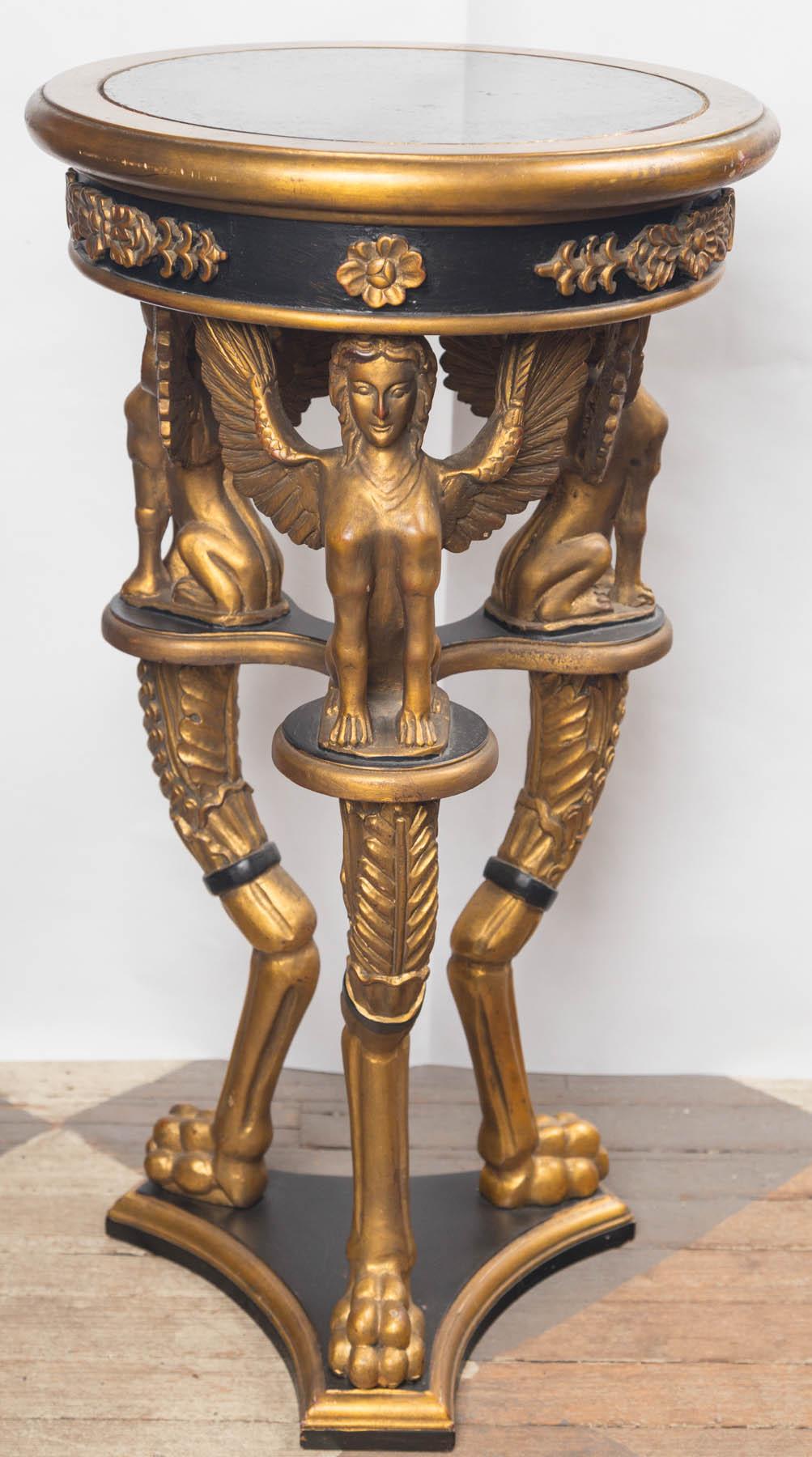 In the Empire style, black and gold surface. Black glass inset top with 15 inches of usable surface. Gold colored appliques around the edge of the top. Three winged female figures with animal legs and paws. Below are three carved incurving legs