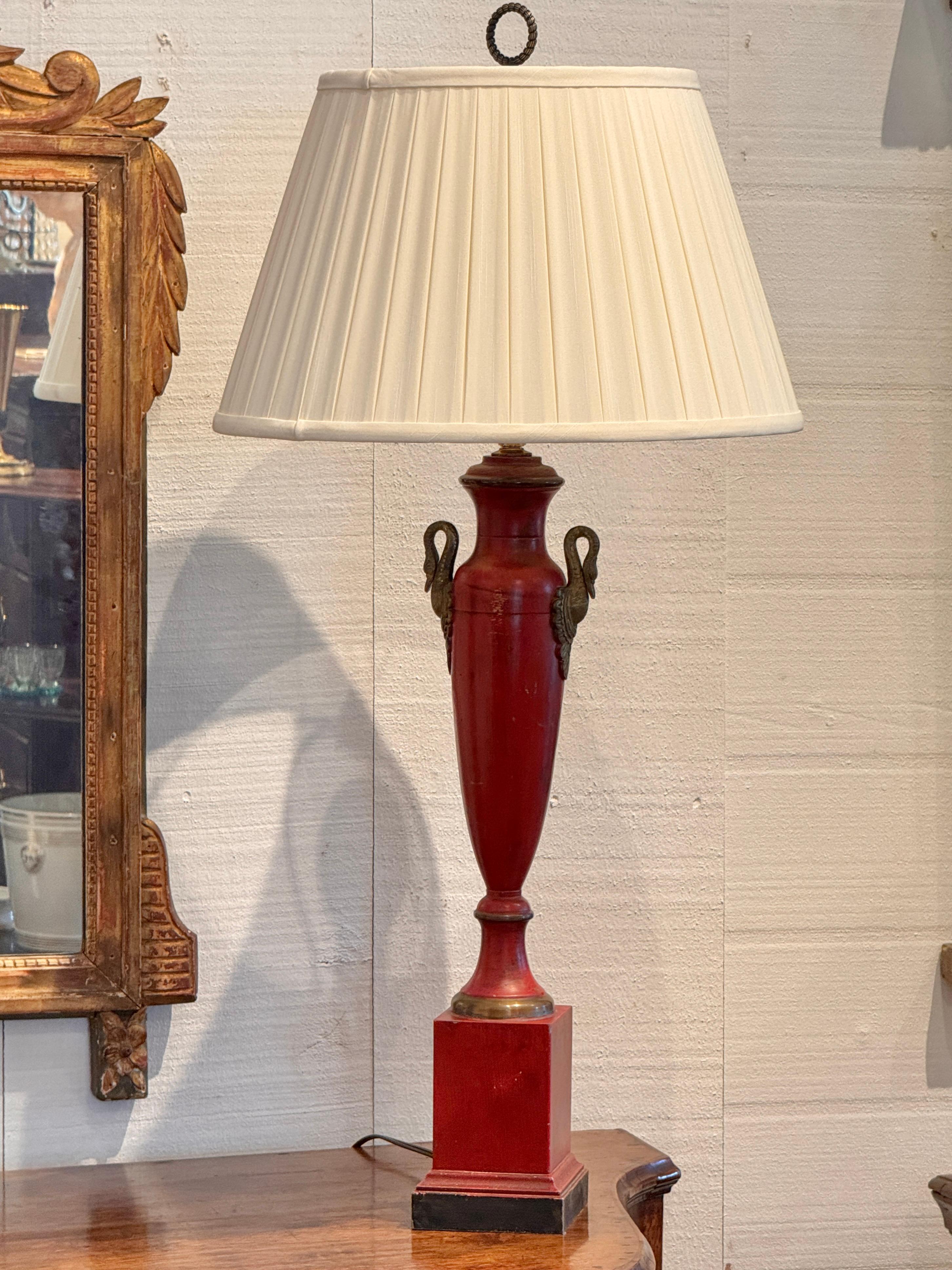 Painted in deep oxblood red with gilt accents, this Empire style tole lamp features swan-form mounts at the neck and stands on a square plinth base with black-painted trim. The swan motif and classic urn silhouette evoke early 19th century French