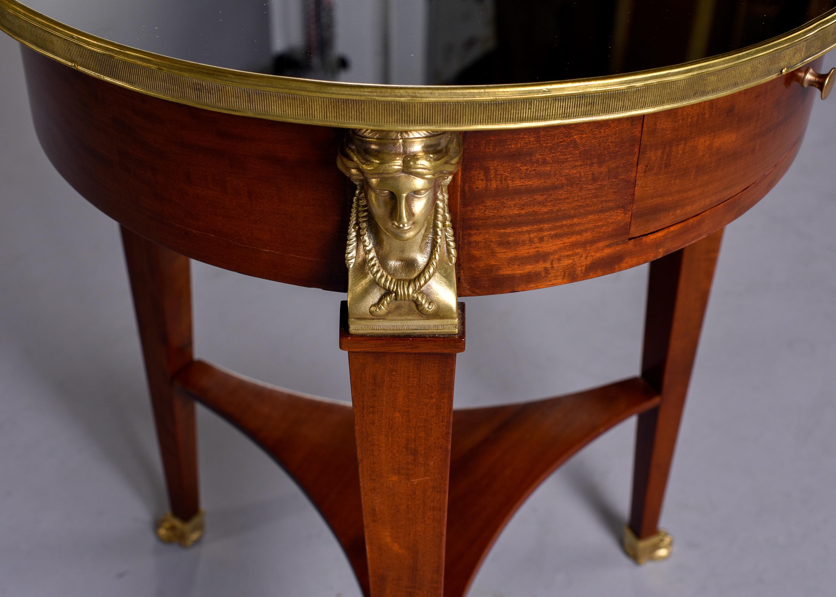 Circa 1920s three legged French Empire style walnut round side table with mirrored top, brass rim and feet, tapered legs topped with sculptural busts in brass and a small pull out drawer. Unknown maker. Very good antique condition.
