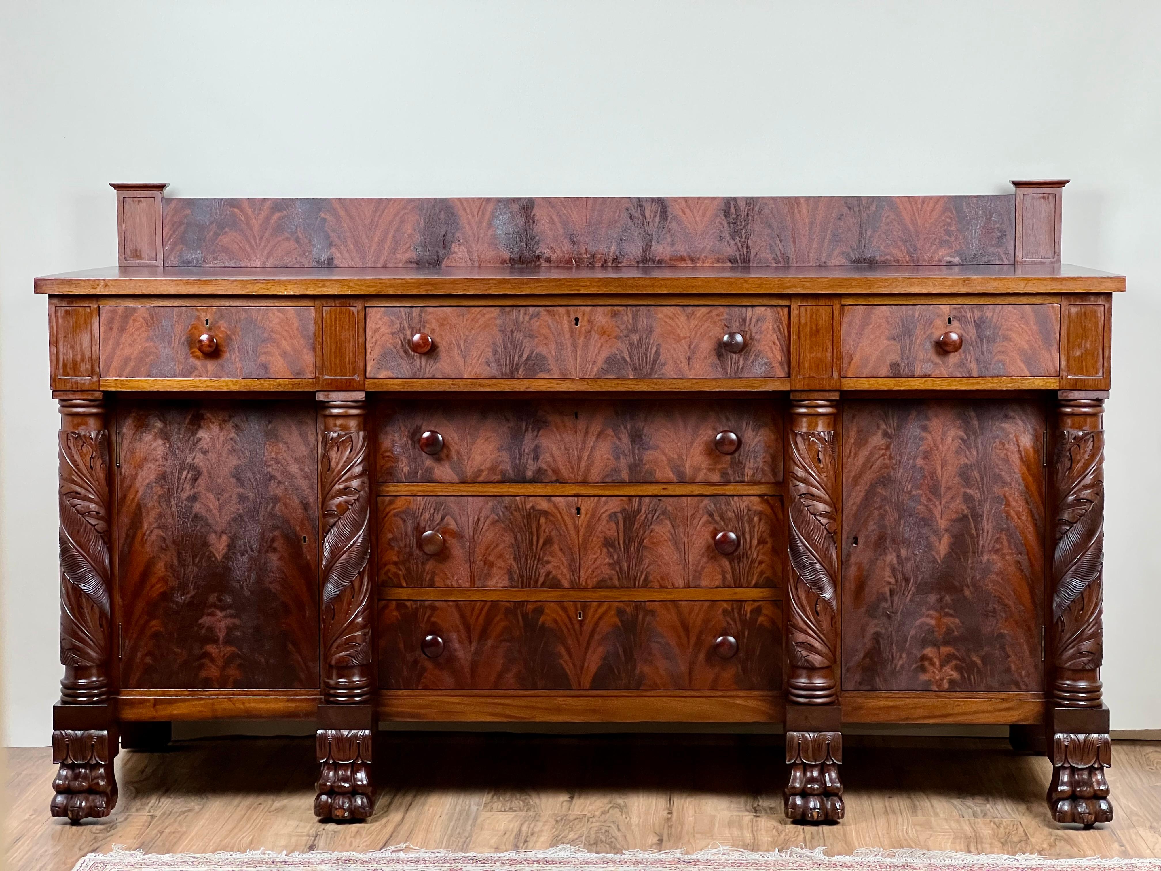 This magnificent American Empire sideboard, crafted in rich mahogany, exemplifies the grandeur and confidence of early 19th-century design. Its bold proportions are enhanced by deeply carved acanthus leaf columns and paw feet, hallmark features of
