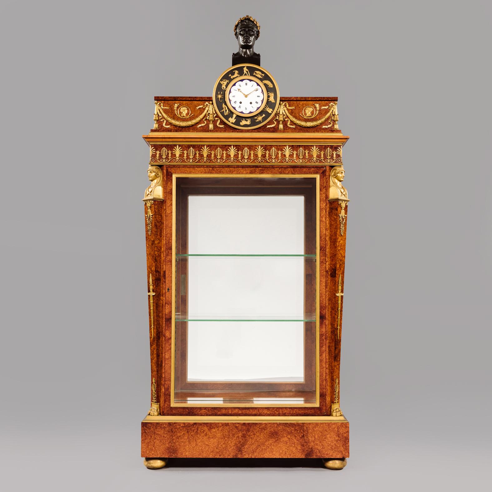 A very fine Amboyna wood and gilt-bronze empire style vitrine cabinet with a glazed front and sides. The cabinet is mounted all over with superb quality well chiselled gilt-bronze mounts and surmounted by a clock with an enamel dial face surrounded