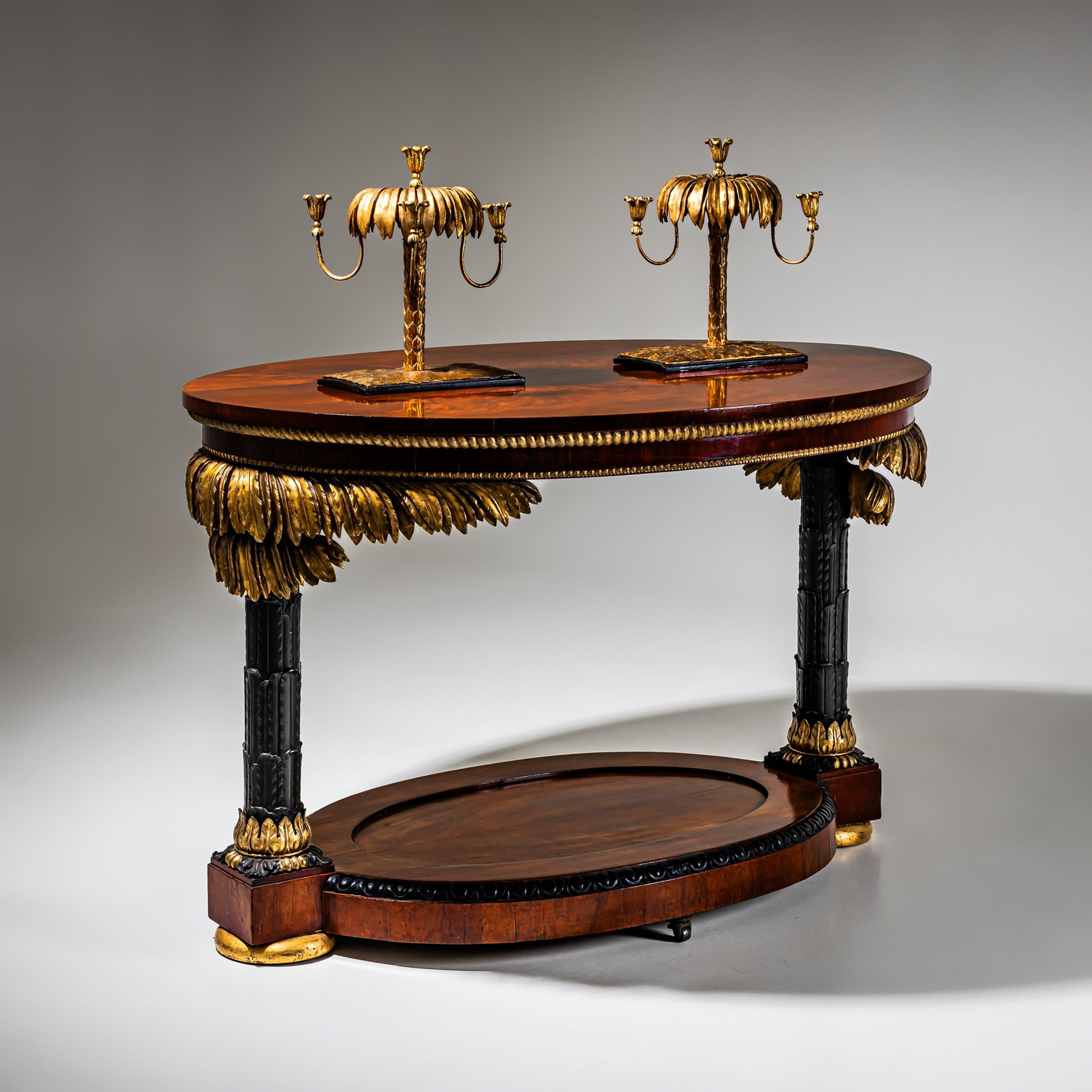 This oval Empire salon table is supported by two columnar legs in the form of stylized palms, partly gilt and partly ebonized. The plinth and the oval top are veneered in mahogany and enriched with kymation and ropework friezes. The distinctive