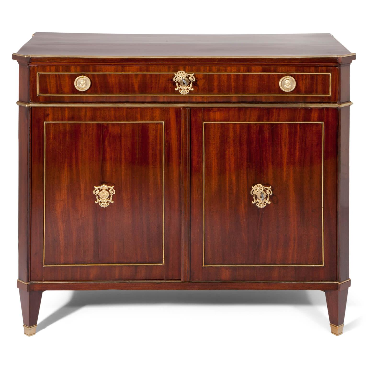 Two-doored cabinet with one top drawer and tapered feet with brass sabots. The corners of the cabinet are slanted. The fillings and edges of the cabinet are framed with brass moldings.
