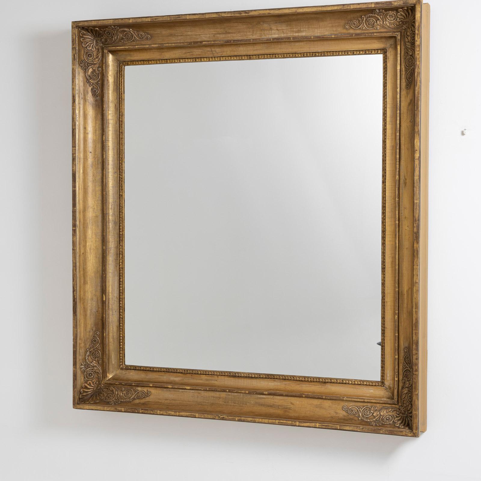 Miroir mural rectangulaire dans un cadre profilé et patiné d'or, avec des motifs décoratifs de feuilles d'acanthe et de rinceaux dans les angles.