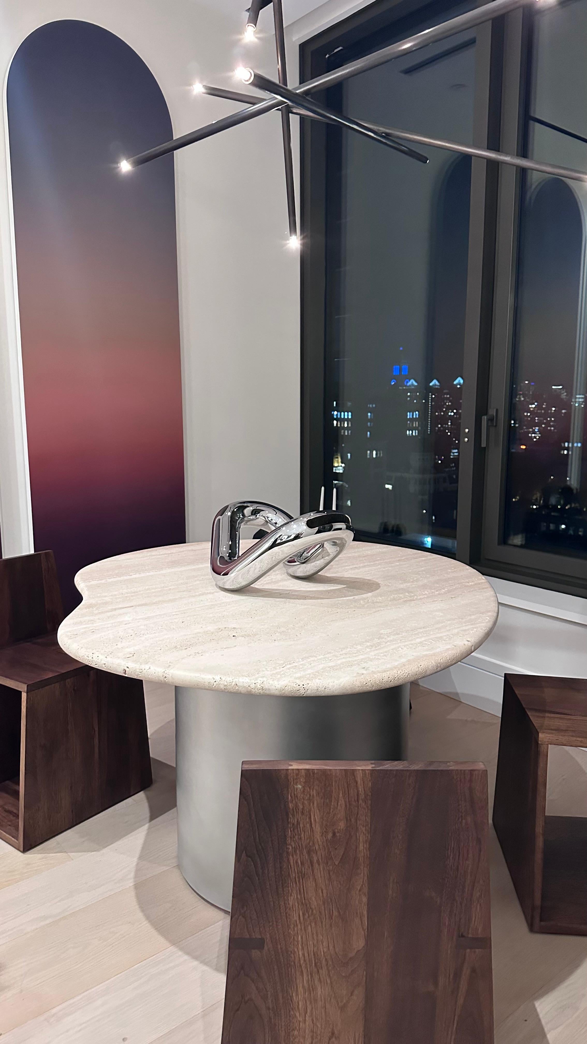 La table Ena présente un superbe plateau de forme organique, soigneusement fabriqué en pierre de travertin. Jumelé avec art à un socle en aluminium fini au pinceau et ciré à la main, son design est un mélange intemporel de nature et d'artisanat.
