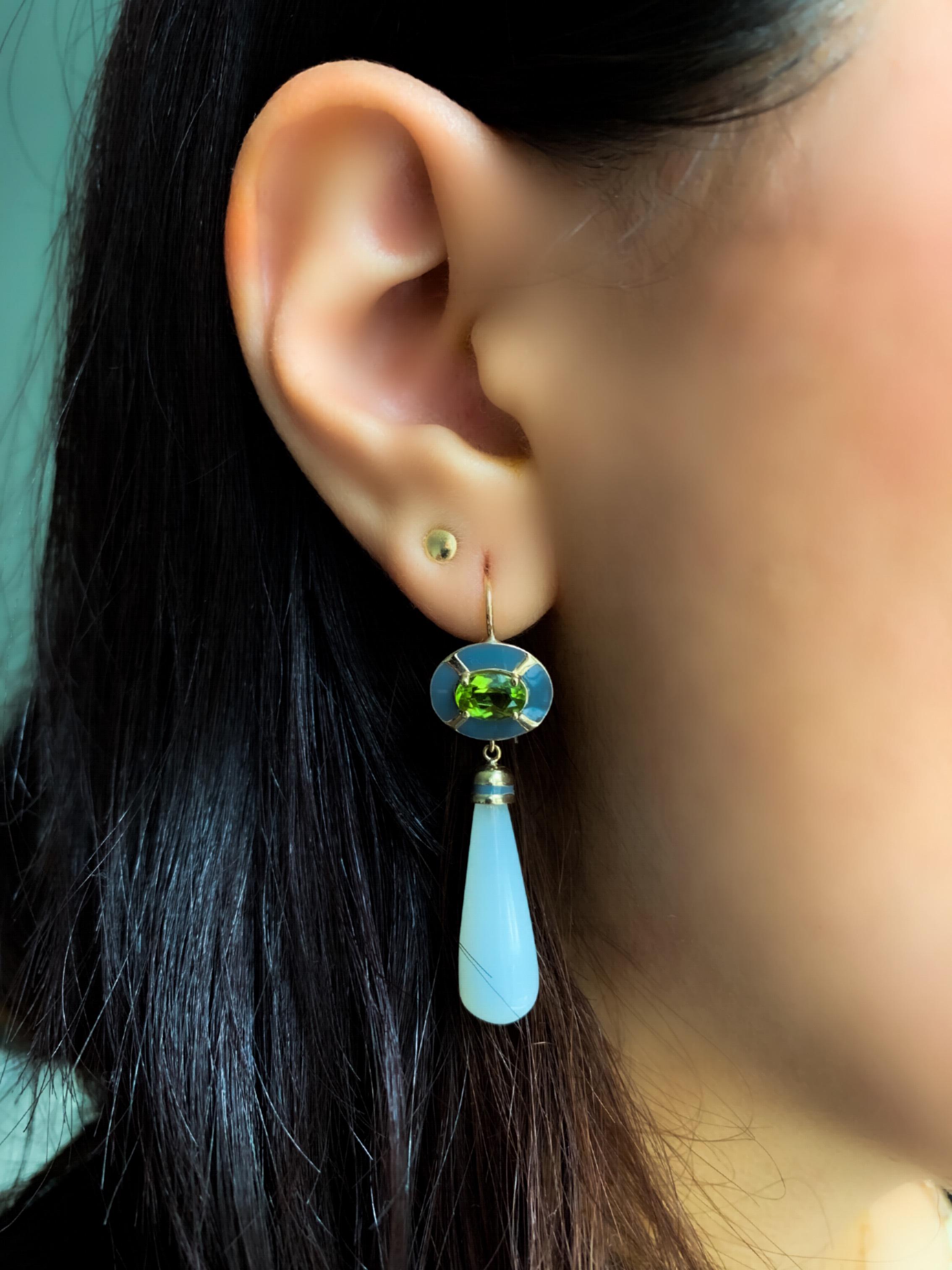 These colorful enamel earrings feature an oval Peridot surrounded by grey enamel.  The design around the peridot is reminiscent of the Art Nouveau period. The Moonstone drop is capped in gold with a stripe of grey enamel.  
Materials: 14 karat gold,